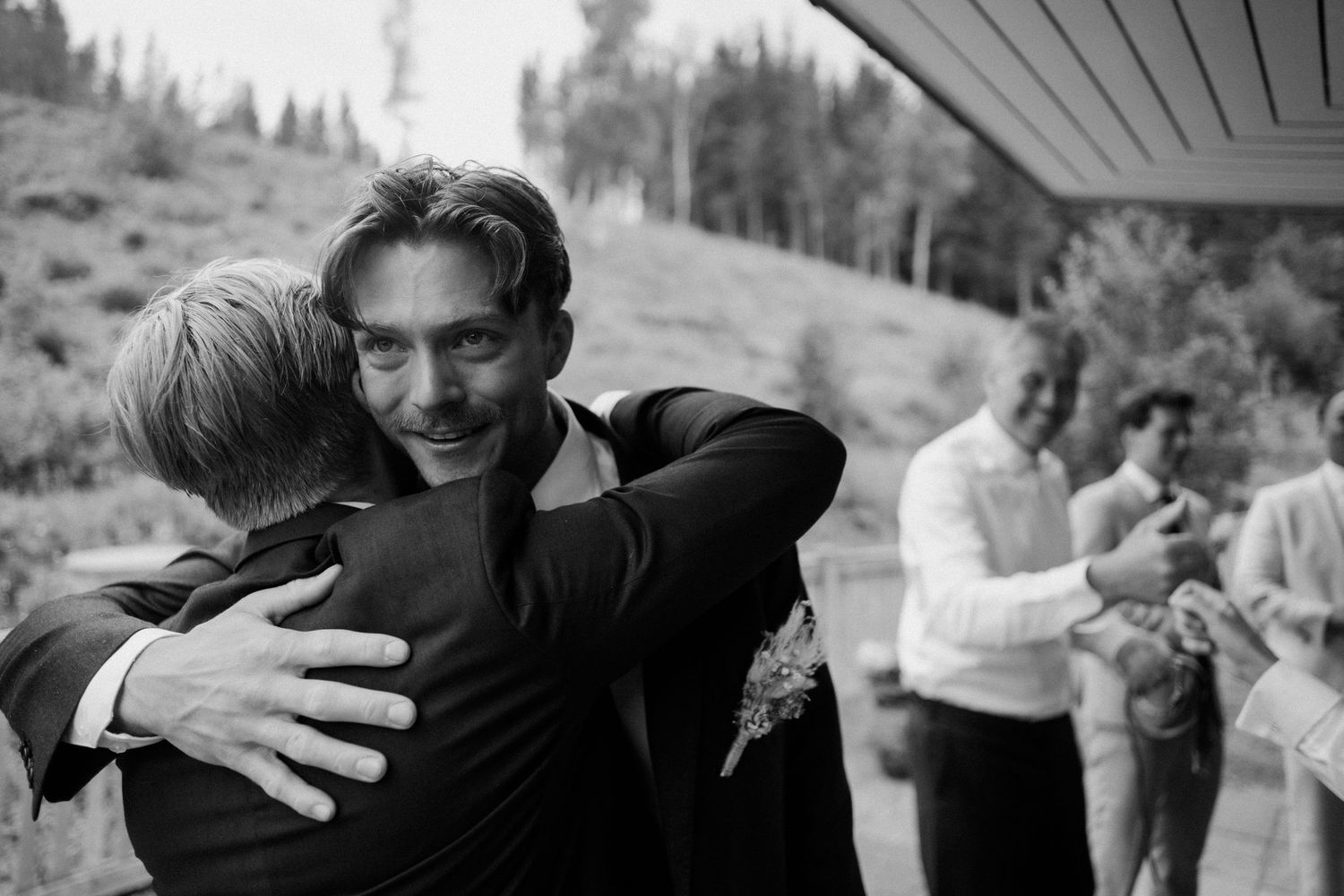 Black and white photo of groom sharing emotional hug with guest at wedding celebration.