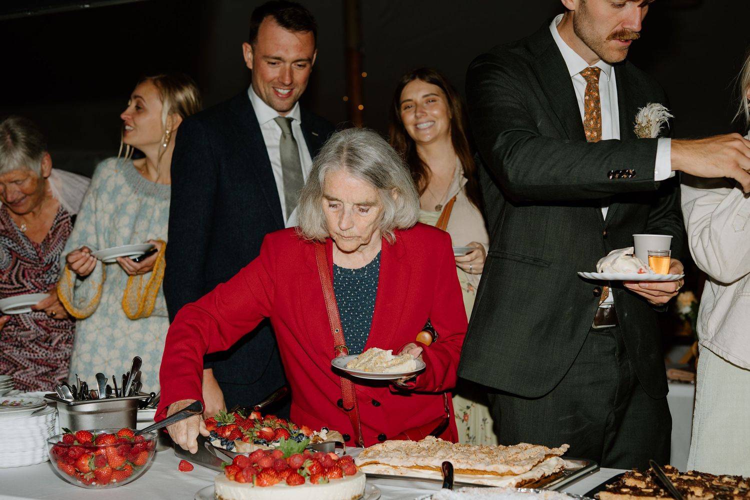 Wedding guests gather around dessert table as woman in red jacket serves herself strawberry cake.