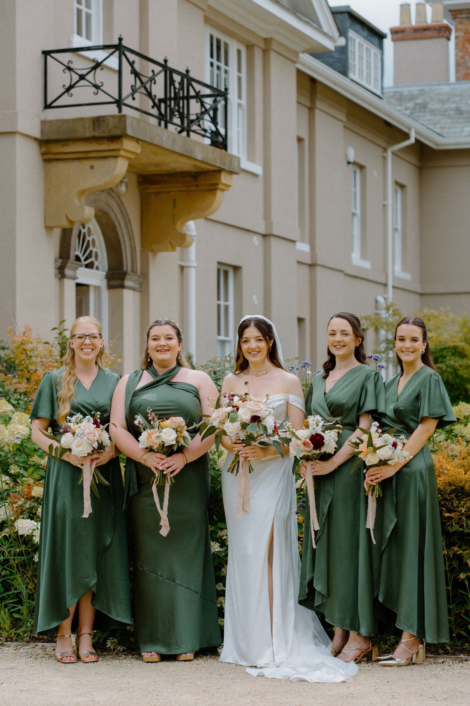 Bride in white gown with four bridesmaids in emerald green dresses holding bouquets outside manor house.
