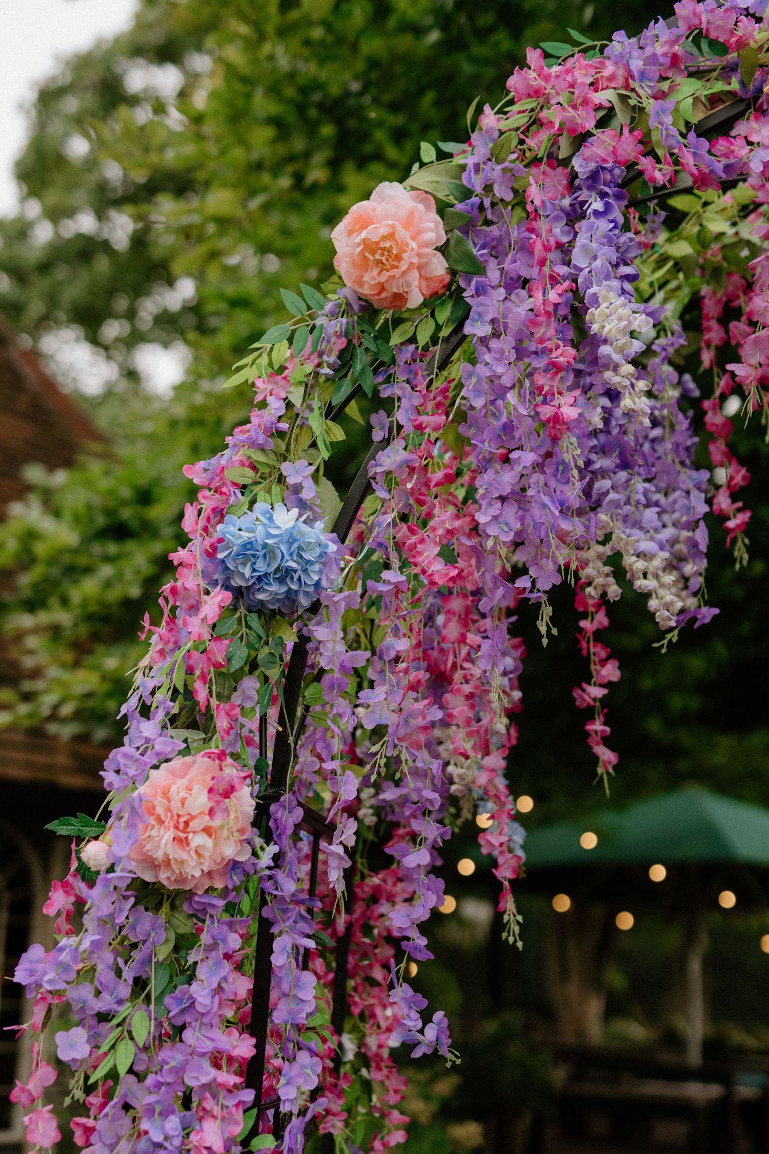 A stunning floral arch adorned with cascading purple wisteria, pink roses, and hydrangeas for a wedding.