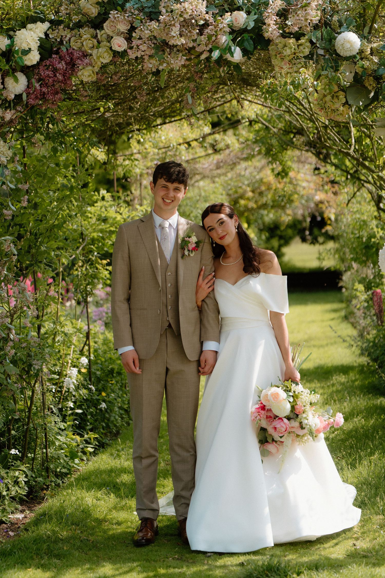 Bride and groom posing under a floral arch in a lush green garden on their wedding day.