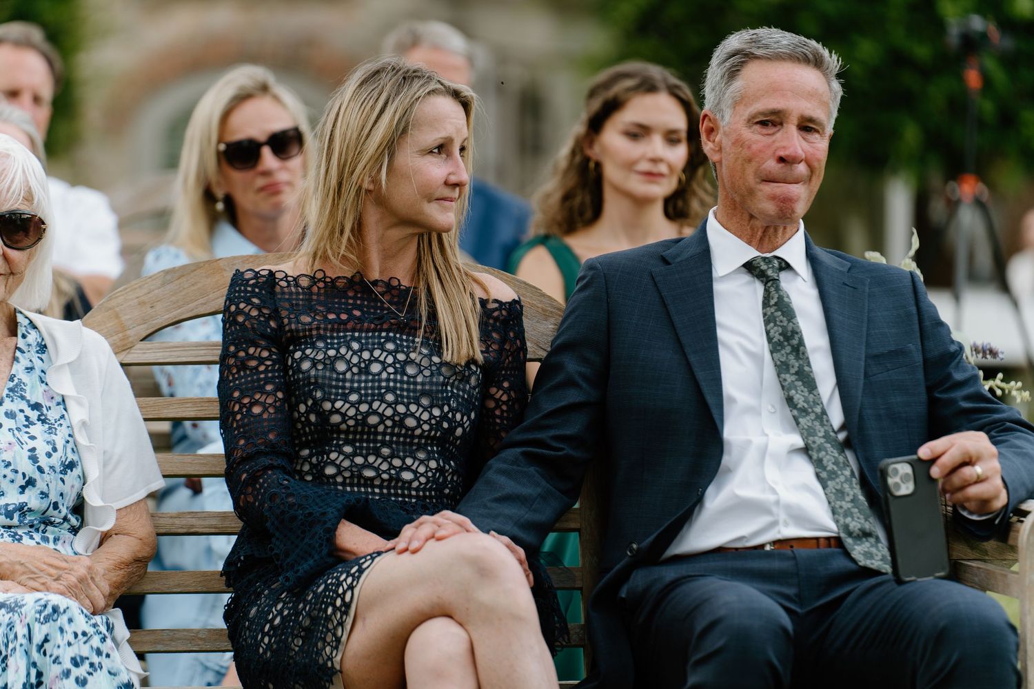 Wedding guests seated outdoors, woman in black lace dress and man in suit watching ceremony.