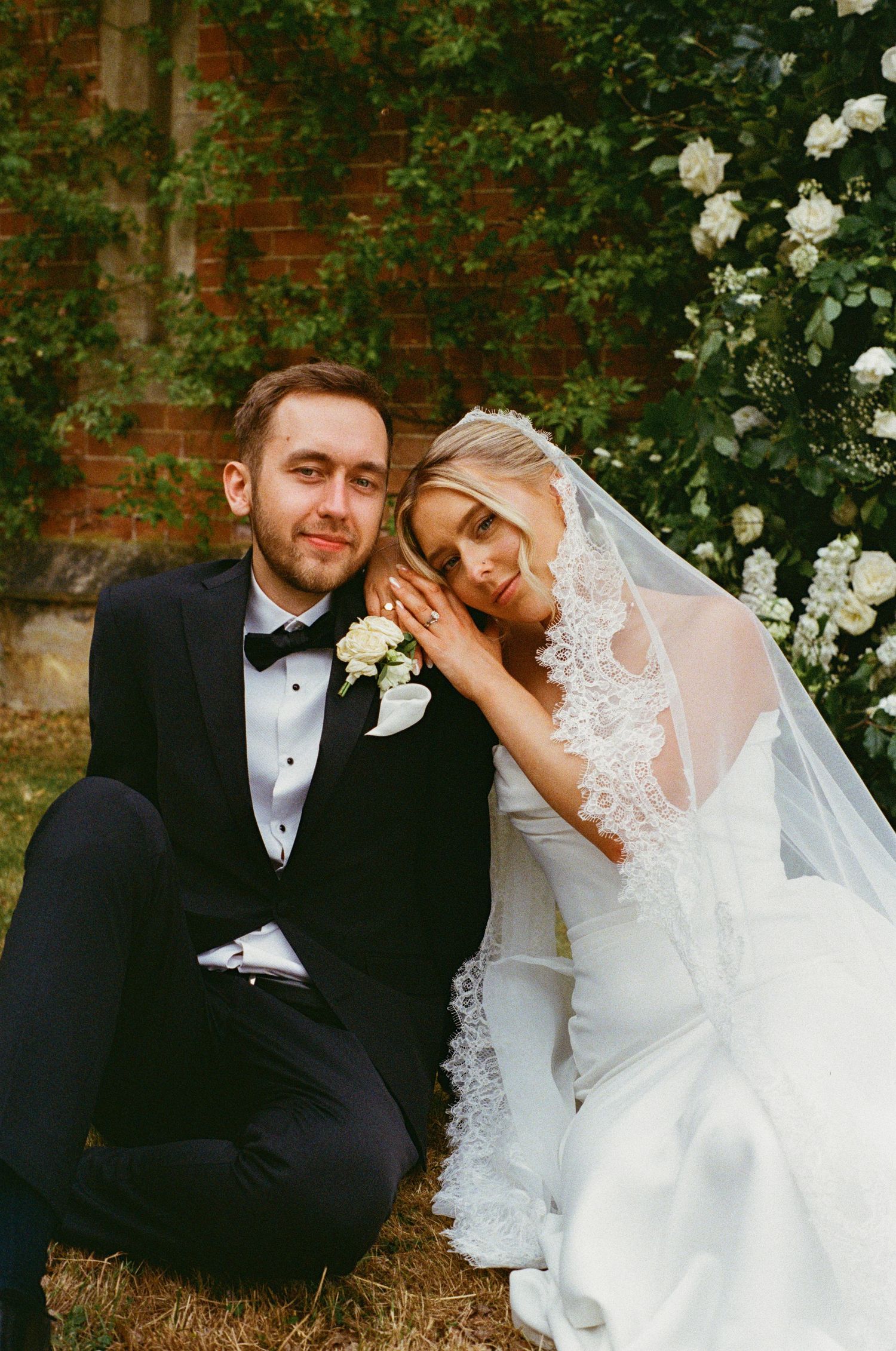 Bride in lace veil rests head on groom's shoulder as they sit together beside white floral arch.