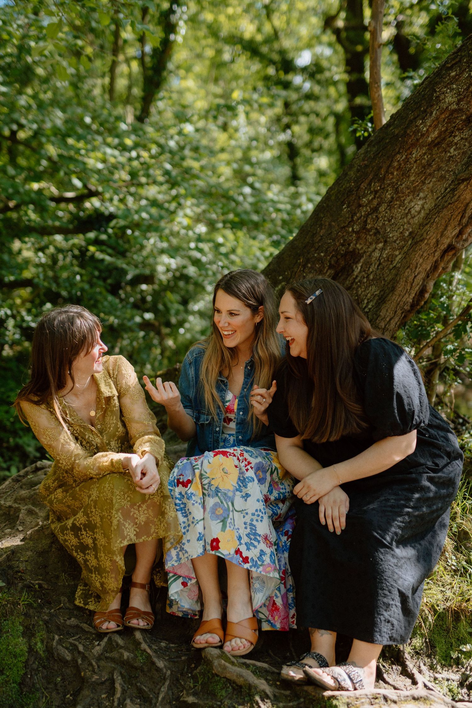 Three women laughing together while sitting on a rock in a sunlit forest setting.