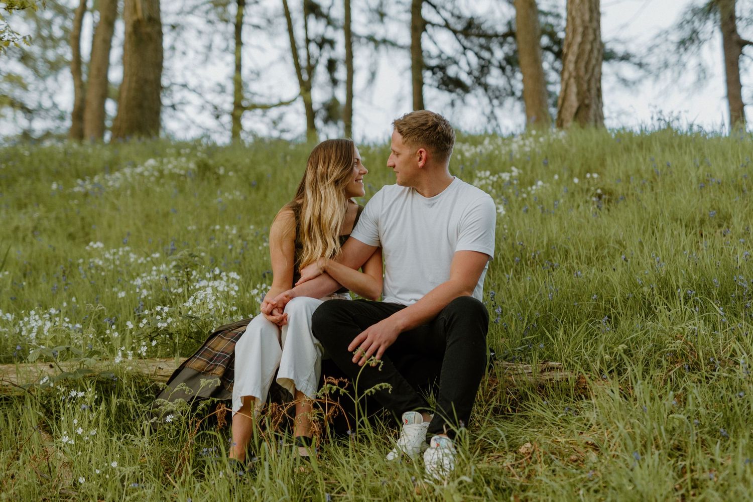 Couple sitting on forest floor surrounded by wildflowers, leaning in for a kiss among tall trees.