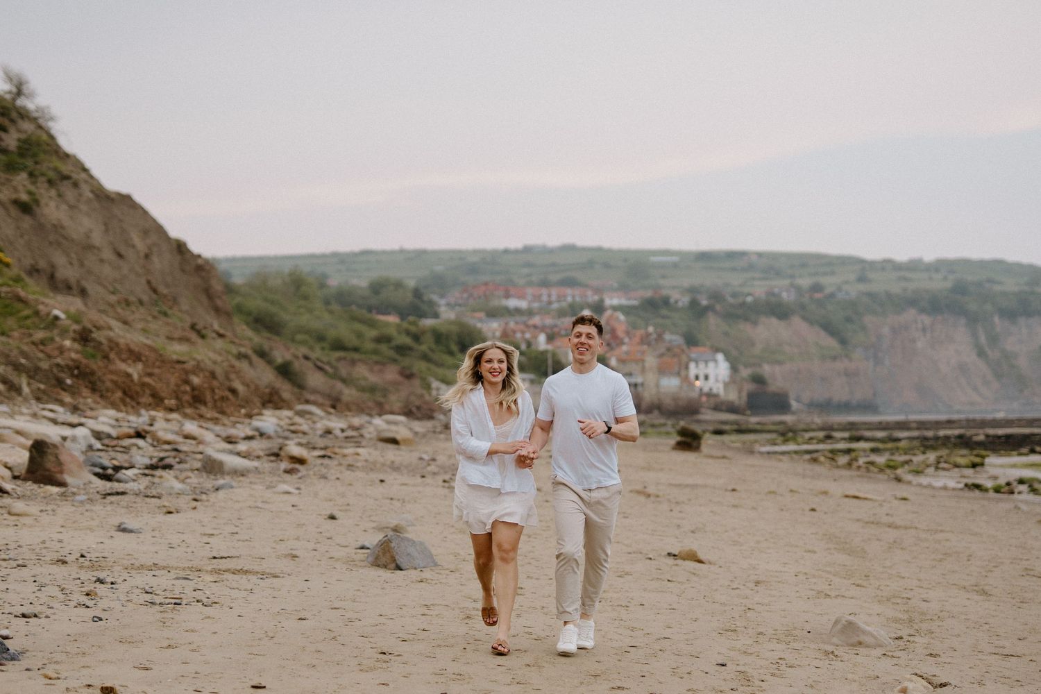 Couple in white outfits running and laughing together on sandy beach with coastal cliffs behind them.