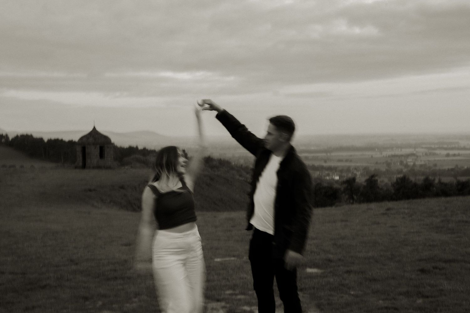 Black and white photo of a couple dancing on a hilltop with dramatic cloudy sky.