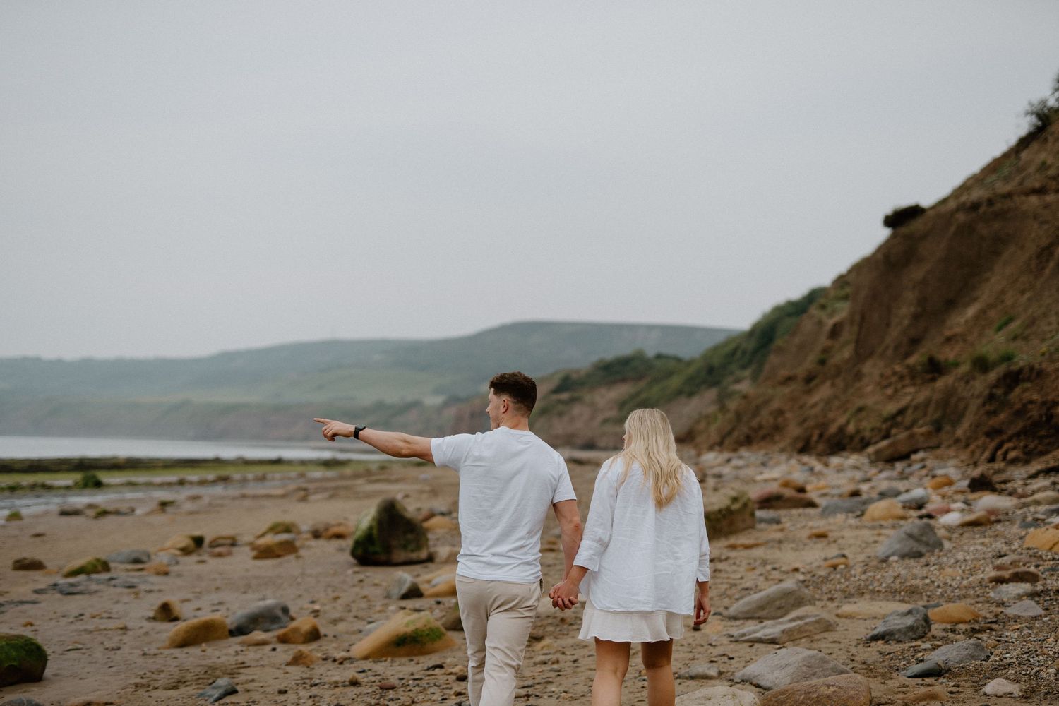 Couple walking hand in hand on a rocky beach, man pointing toward the scenic coastal horizon.