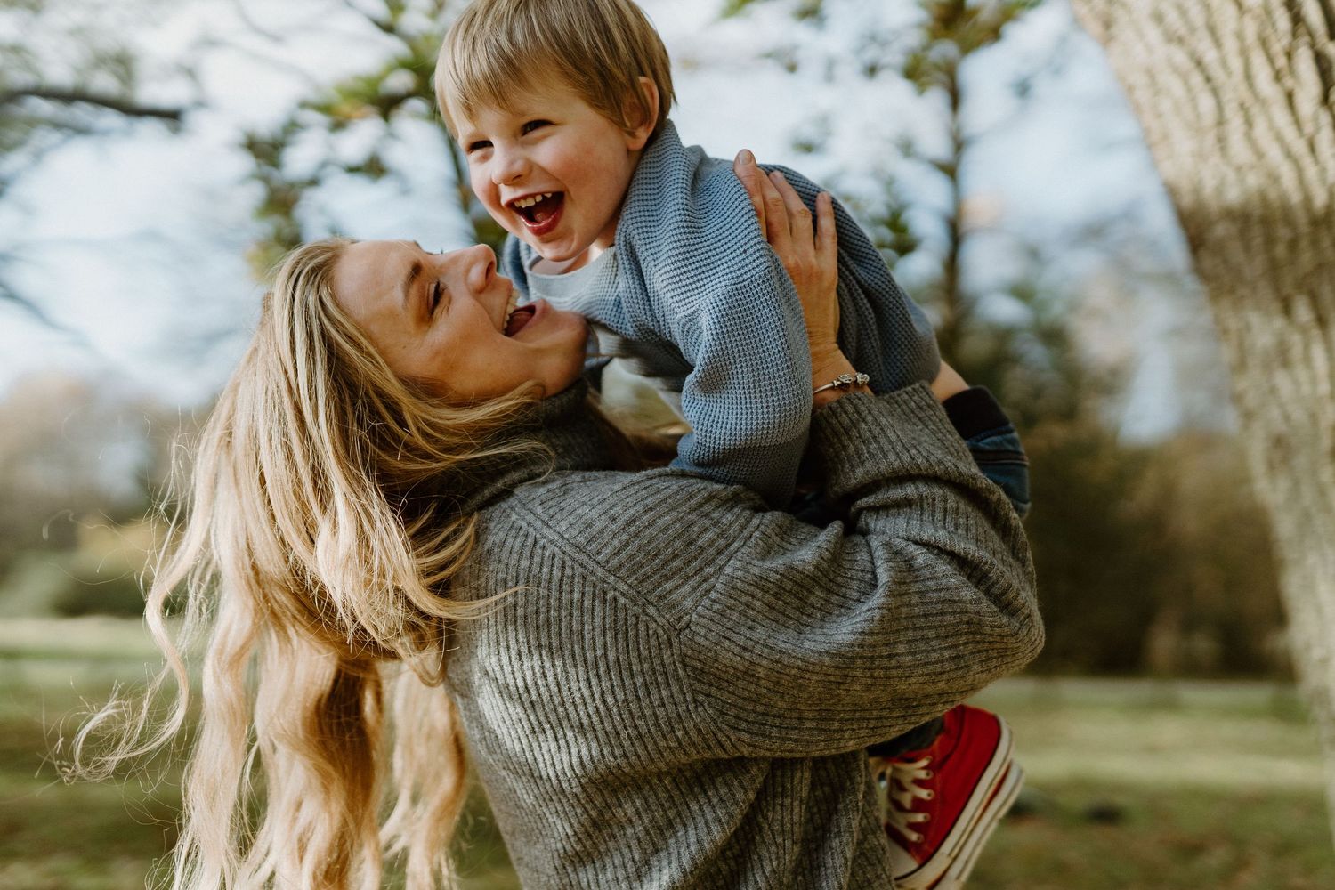 A joyful mother with blonde hair lifts her laughing toddler outdoors in a park setting.
