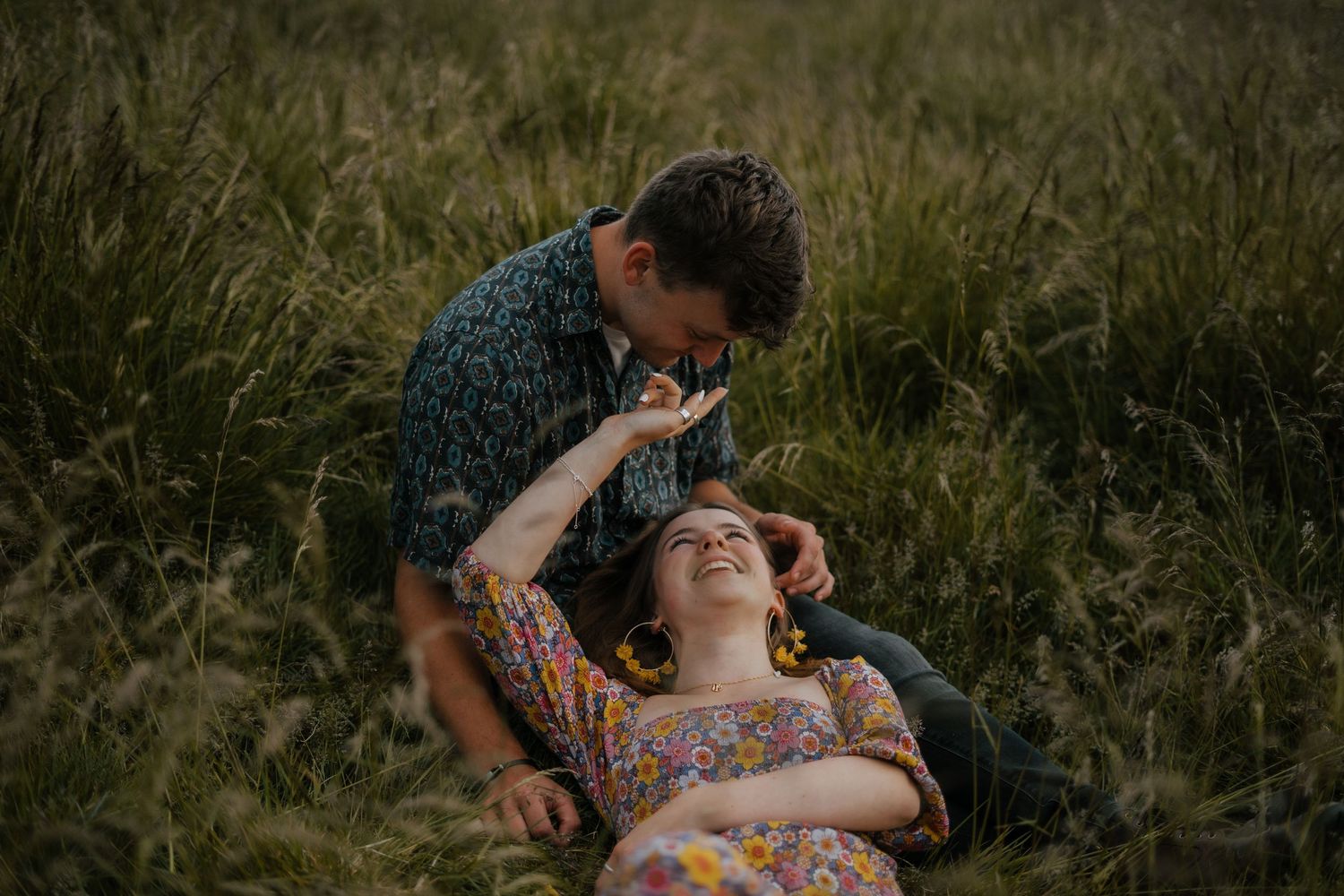 Man gently touching womans hair as she laughs lying in tall grass during couples shoot.