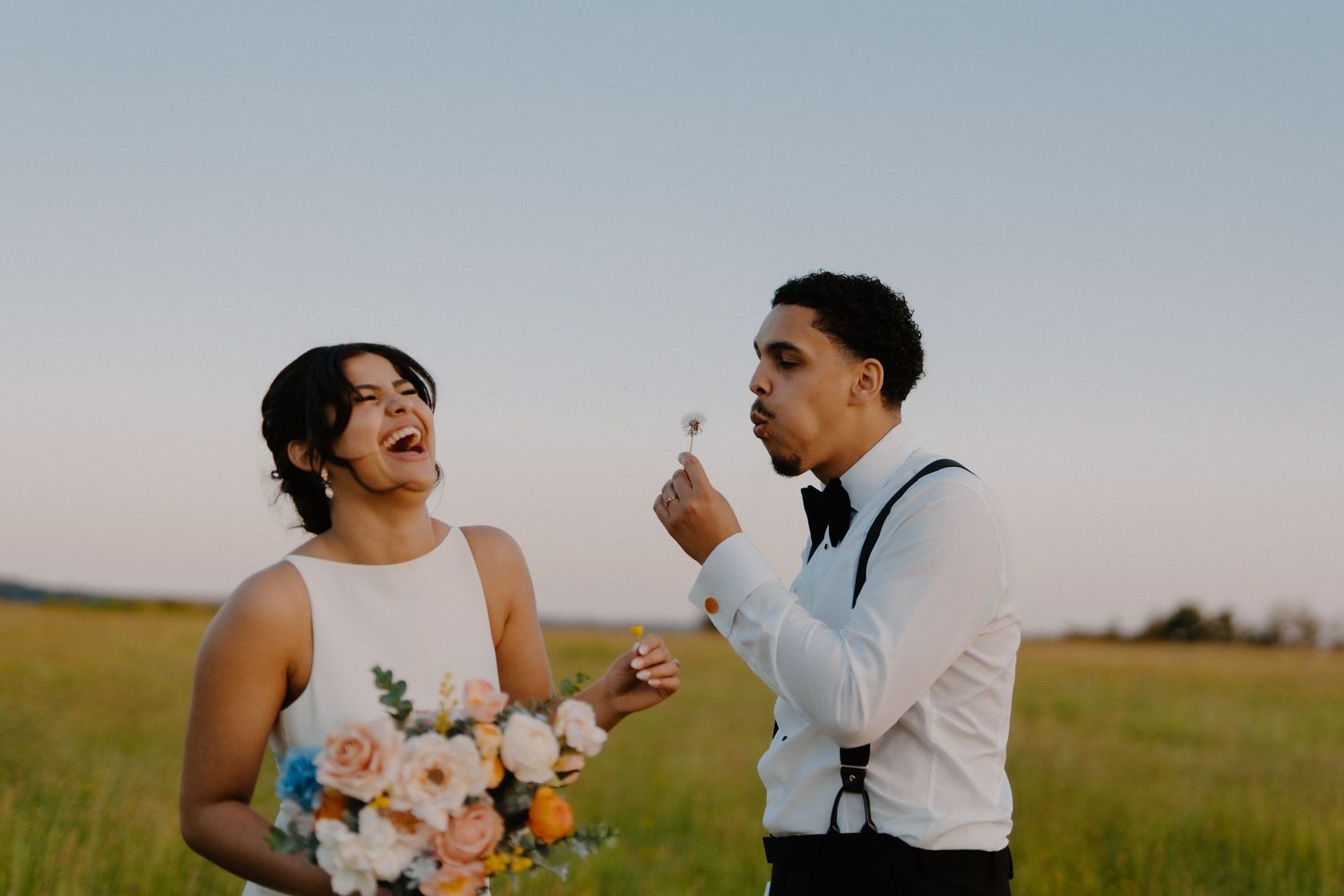 Joyful bride laughing as groom blows a dandelion in a sunlit open field during wedding celebration.