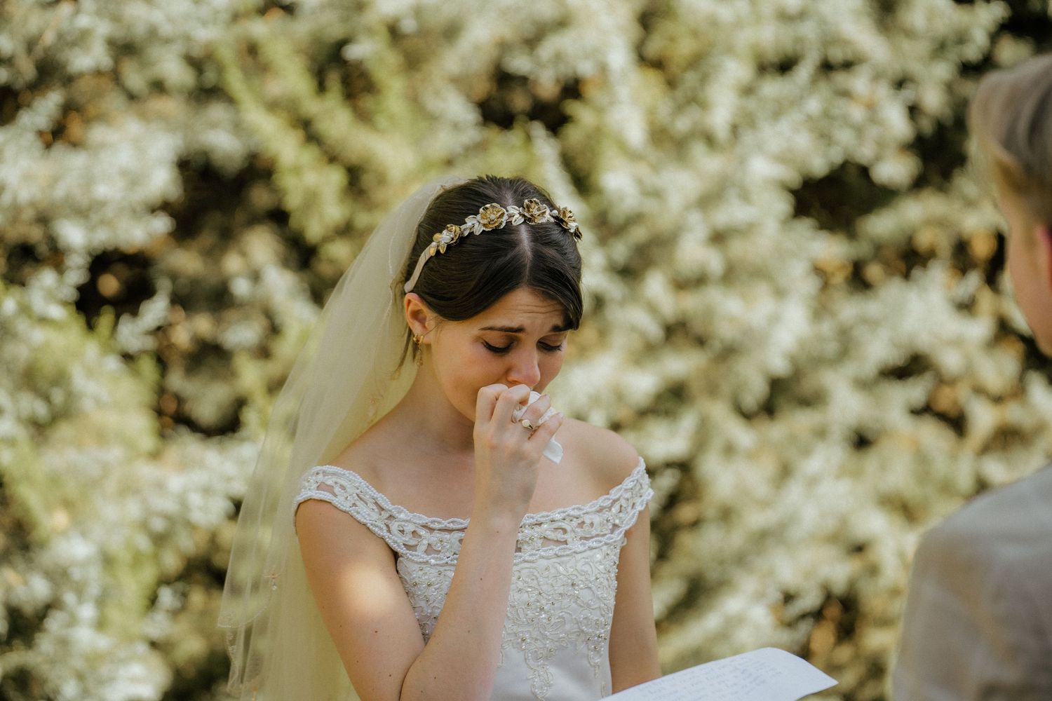 Emotional bride in lace dress and floral headpiece wipes tears during ceremony.