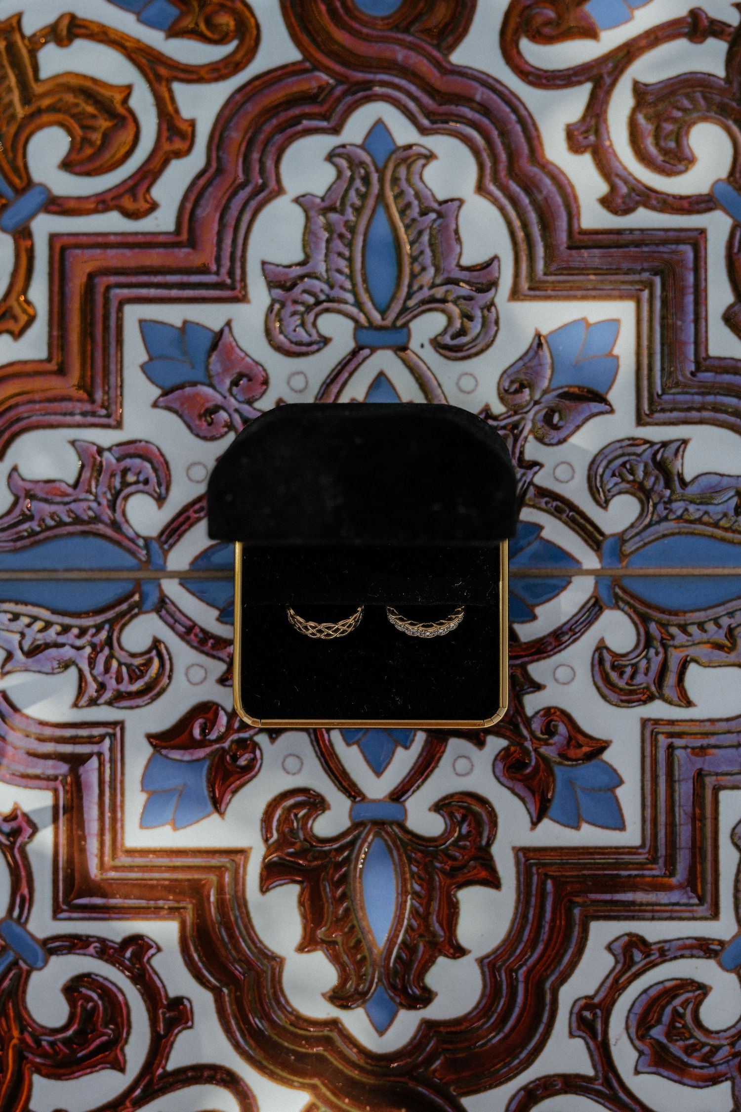 Wedding rings displayed in a black velvet box against ornate blue and brown decorative tile pattern.