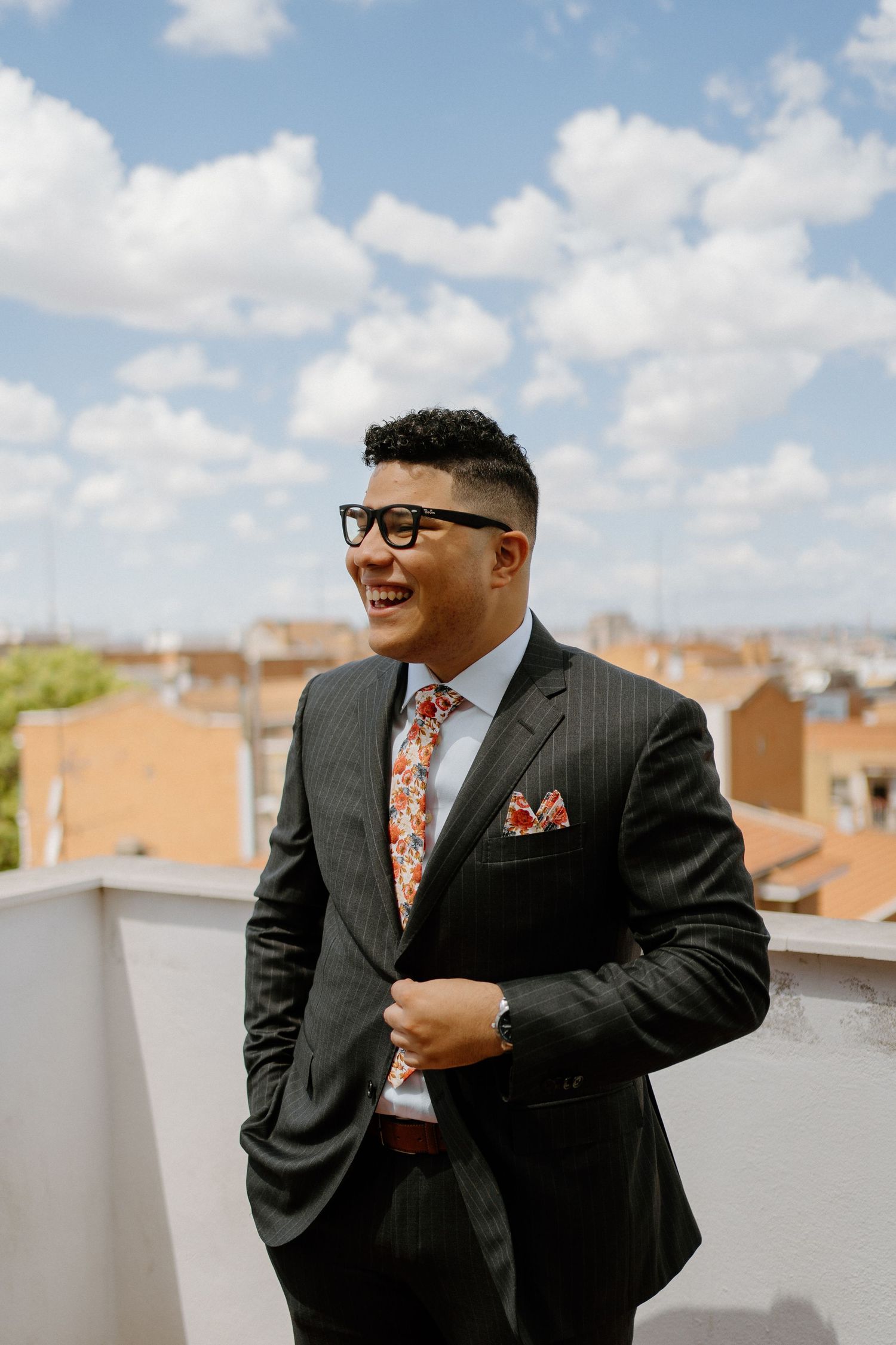Stylish groom in pinstripe suit with floral tie laughs on rooftop under blue cloudy sky.