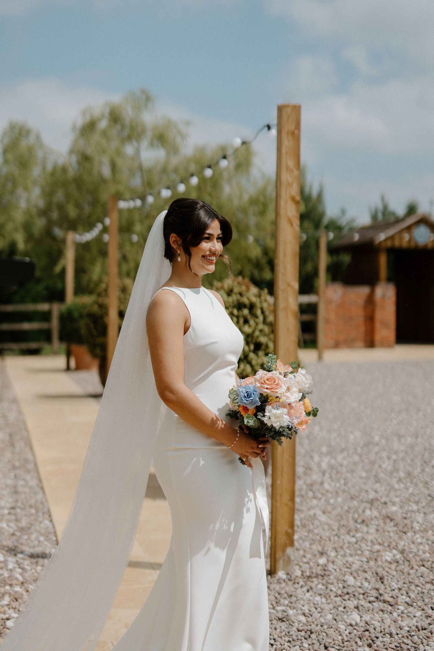 Smiling bride in sleek white gown and veil holding pastel bouquet outdoors on sunny wedding day.