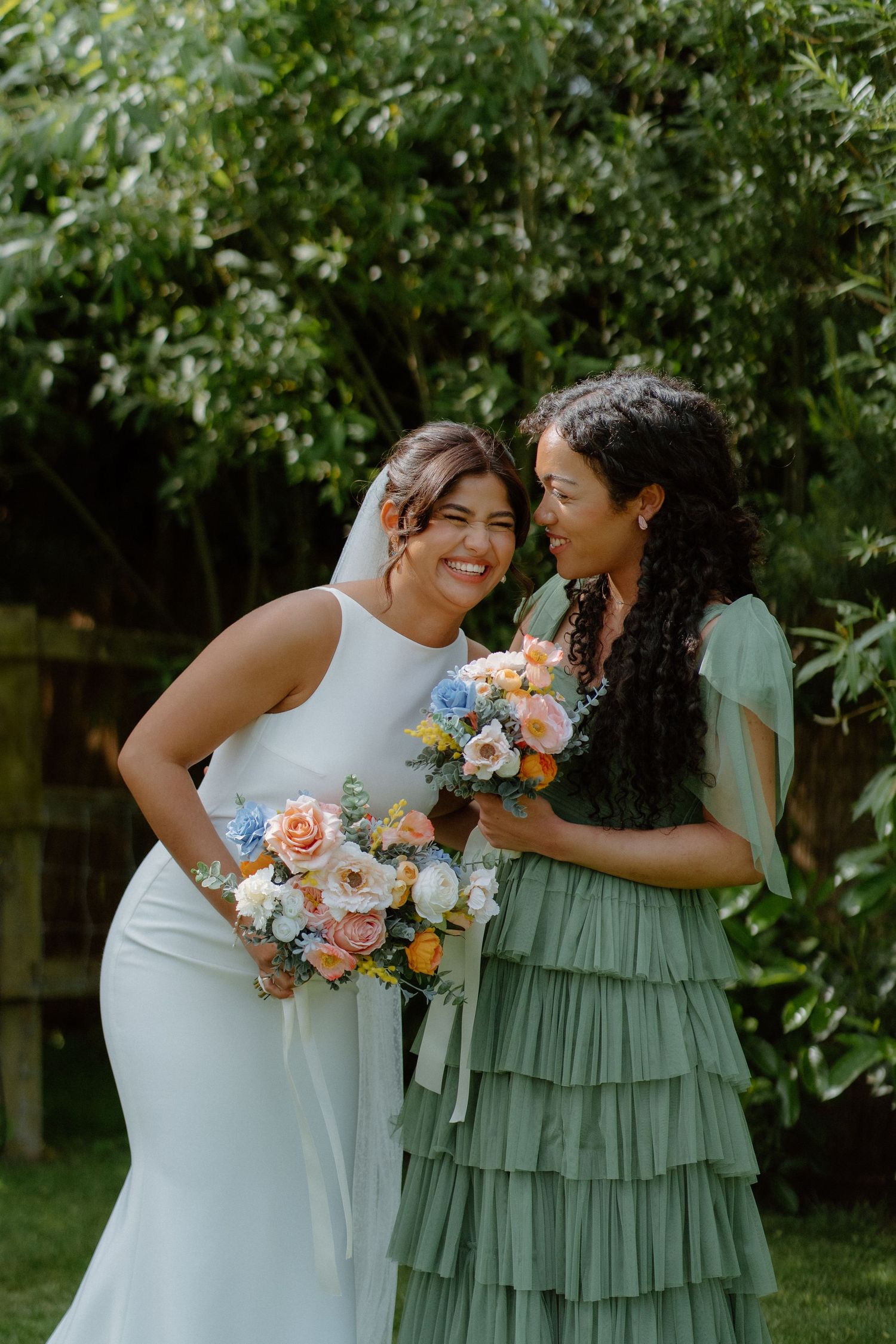 Smiling bride in white halter gown shares joyful moment with bridesmaid in sage green ruffled dress outdoors.