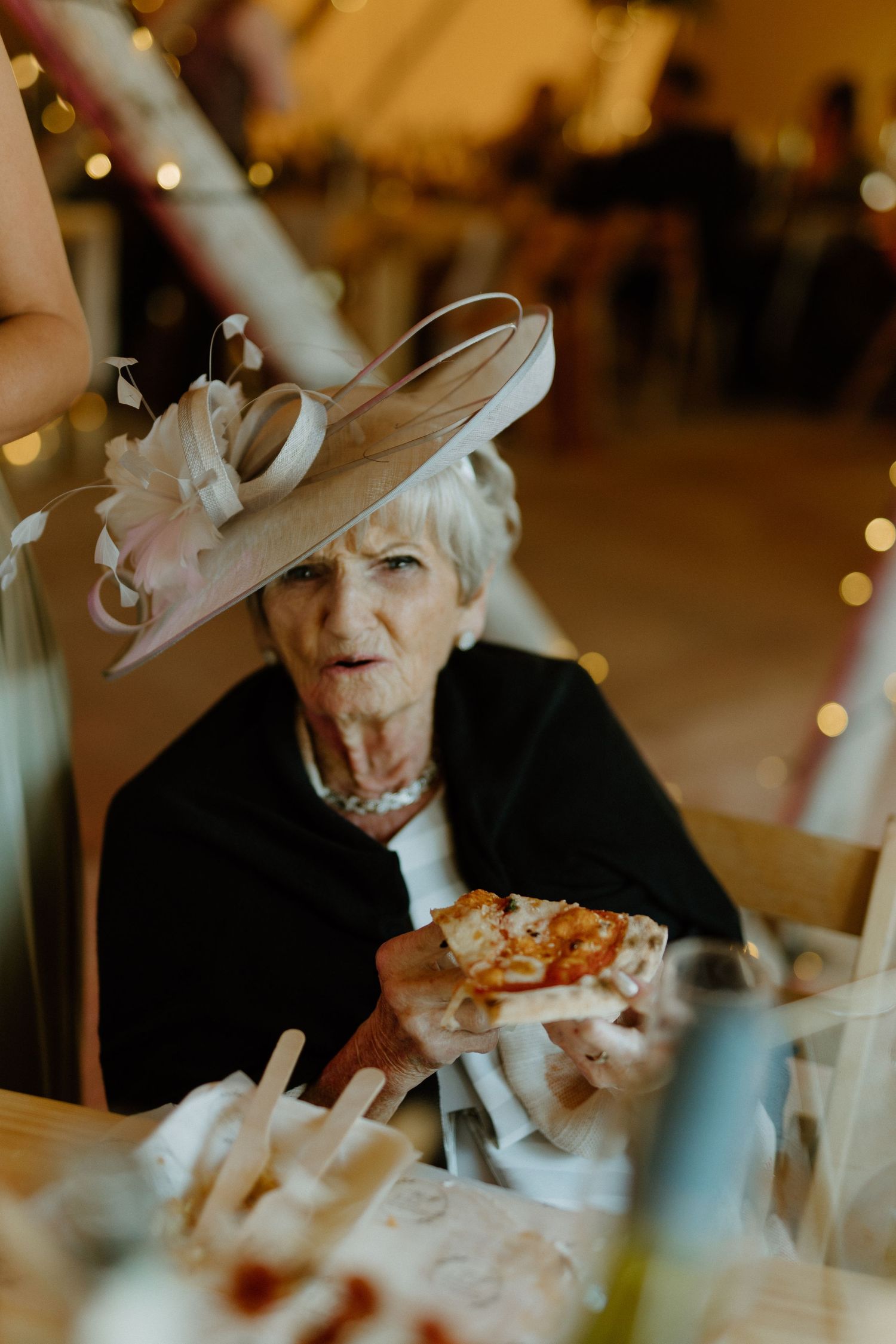 An elegant guest in a wide-brimmed hat and black jacket enjoys food at an evening celebration with twinkling lights.