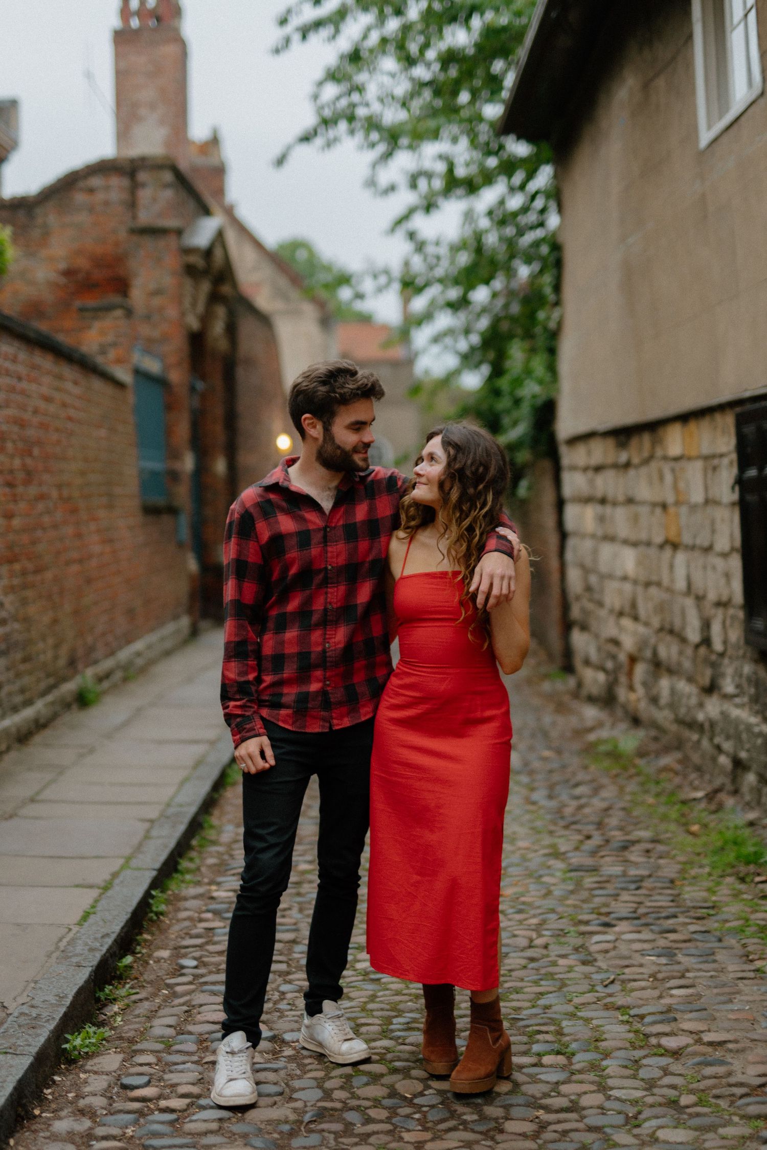 A couple walking on a cobblestone path beside brick buildings, with one wearing red dress and other in plaid flannel.