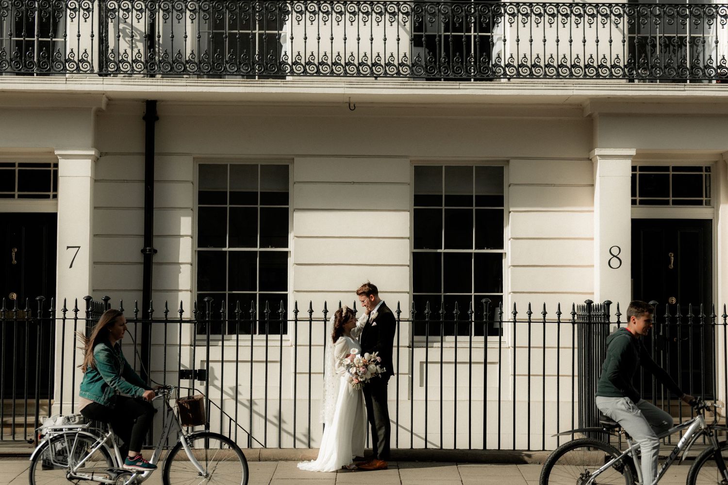A wedding couple poses between numbered doorways while cyclists pass by on a London street.