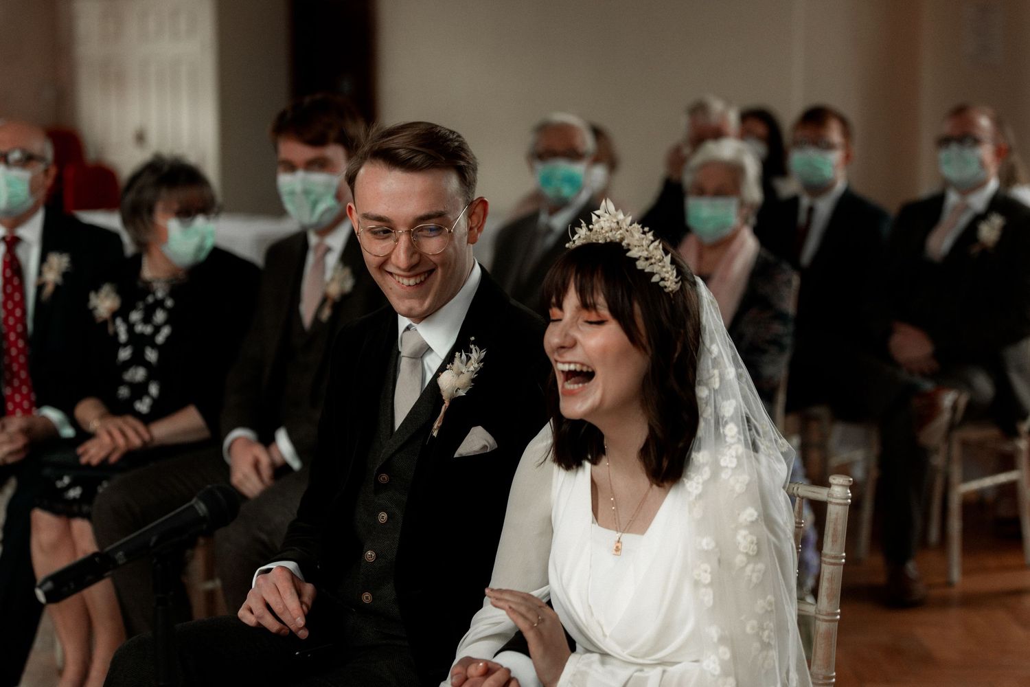 A joyful wedding ceremony moment with guests wearing face masks in the background.