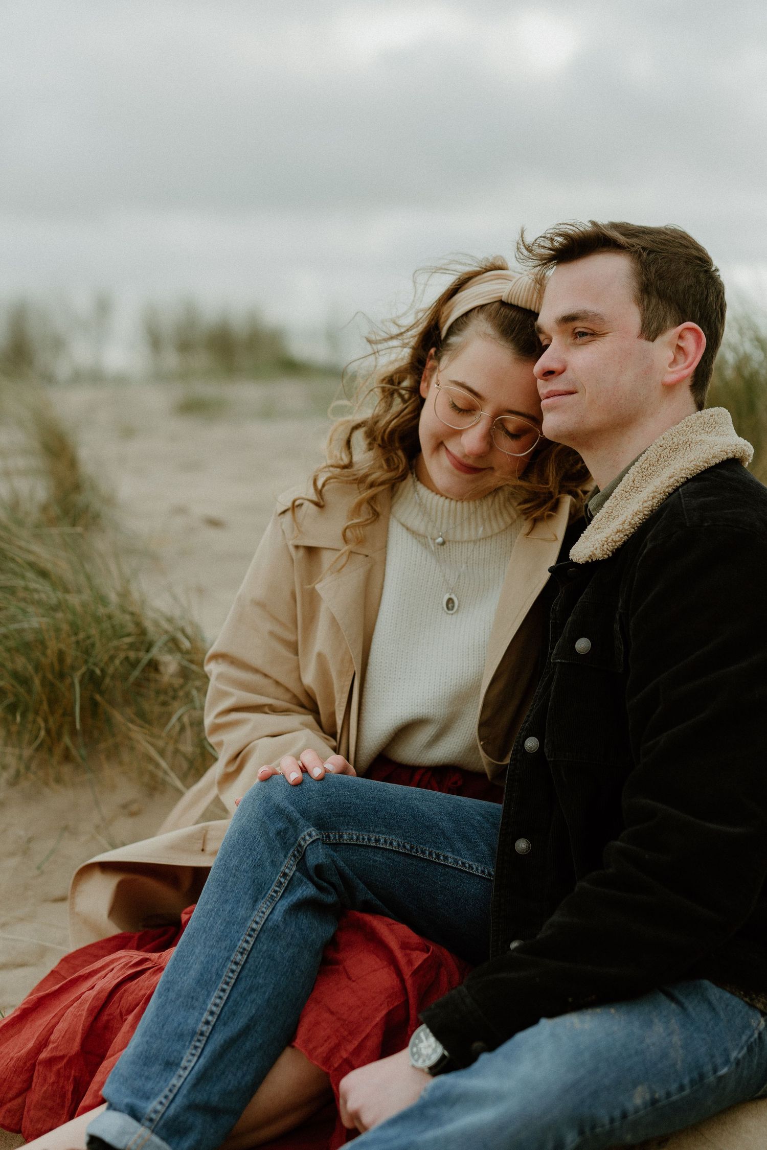 Couple sitting together in sand dunes, woman resting head on mans shoulder tenderly.