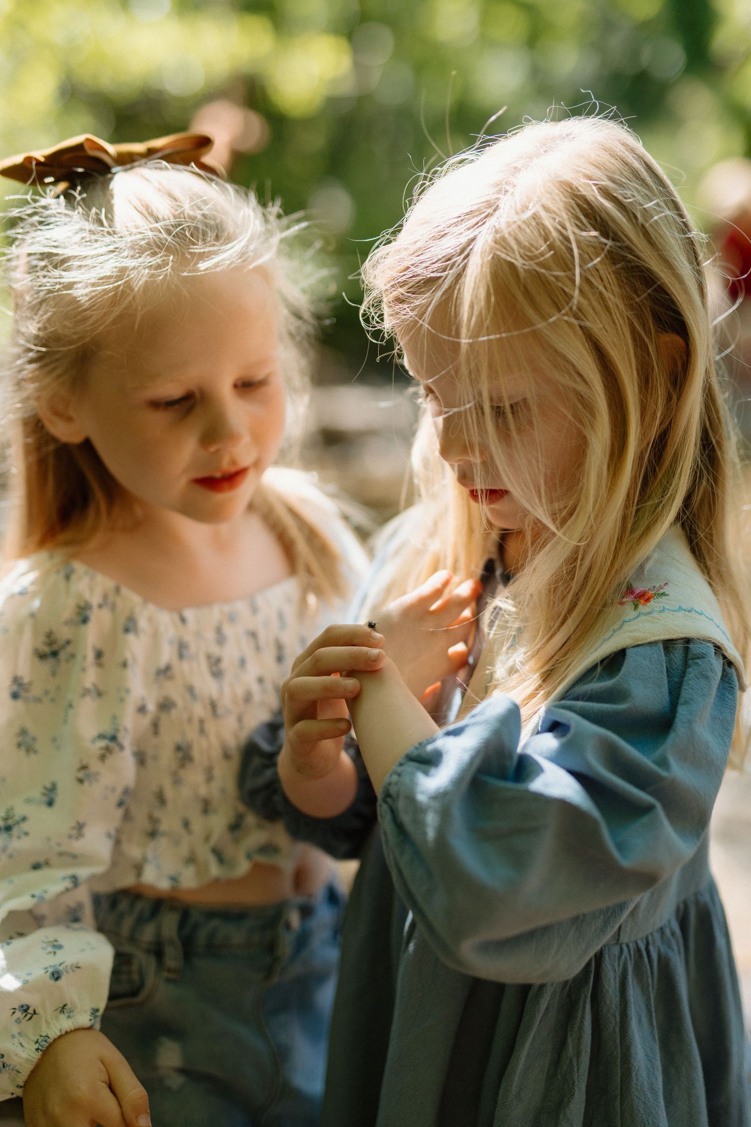 Two young blonde girls in floral dresses sharing a small object in a sunny garden.