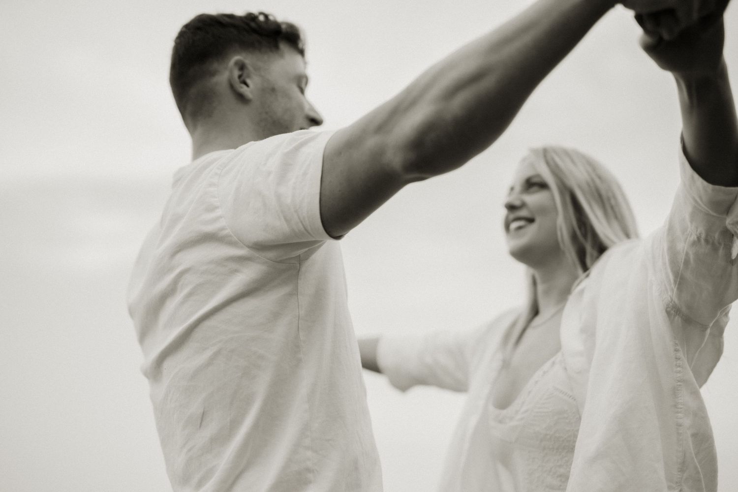 Black and white photo of a joyful couple dancing and holding hands on a beach together.