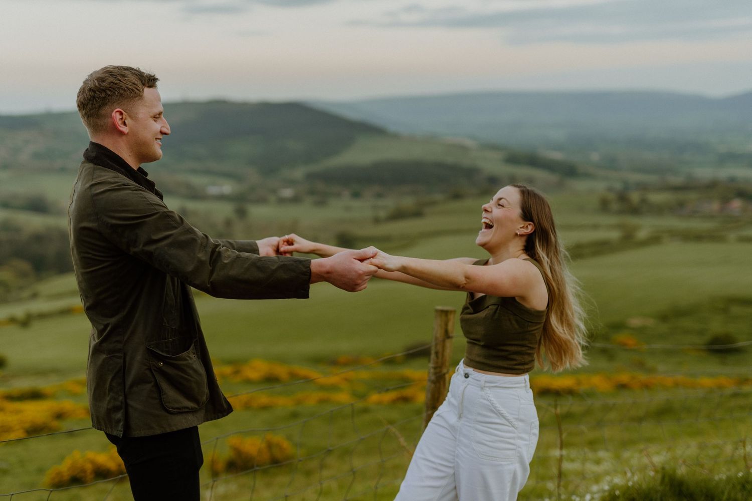 Joyful couple holding hands and spinning in a green meadow with rolling hills.
