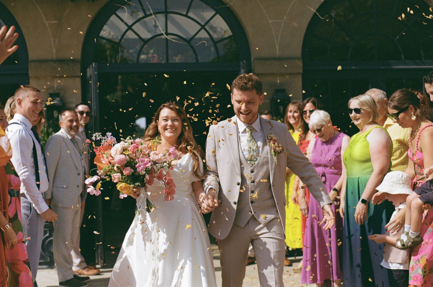 Newlyweds walk through colorful confetti shower surrounded by cheering guests outside elegant building after ceremony.