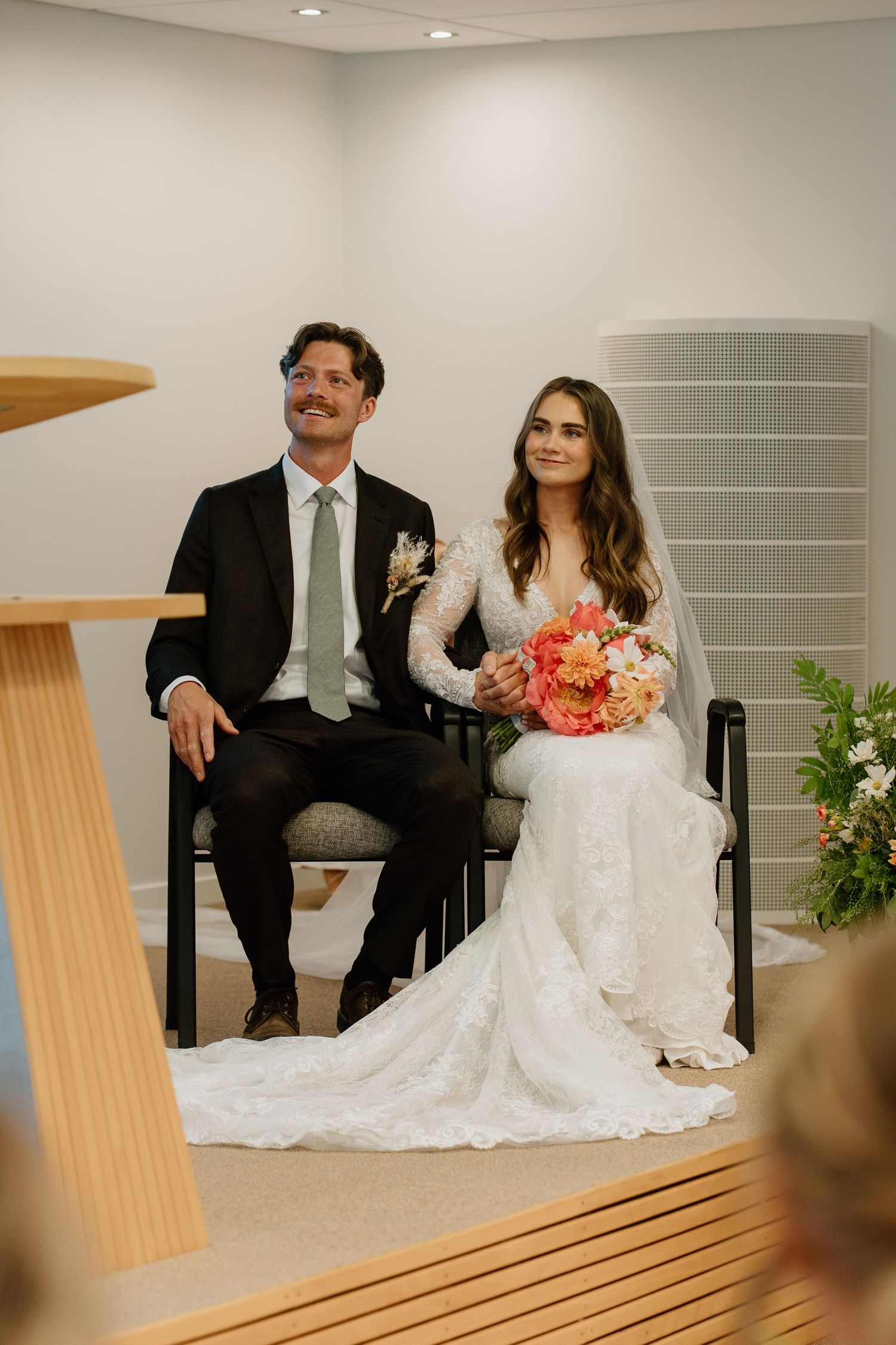 Bride and groom seated at altar during wedding ceremony, bride holding coral floral bouquet.