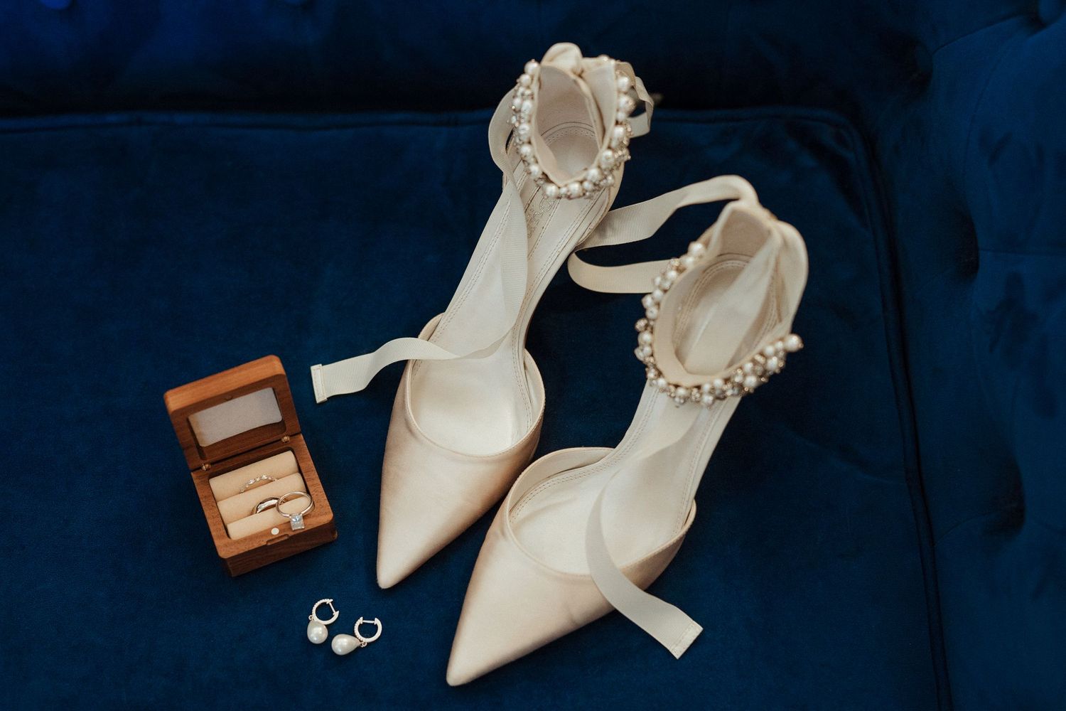White pearl-adorned bridal heels with ring box and earrings displayed on blue velvet surface.