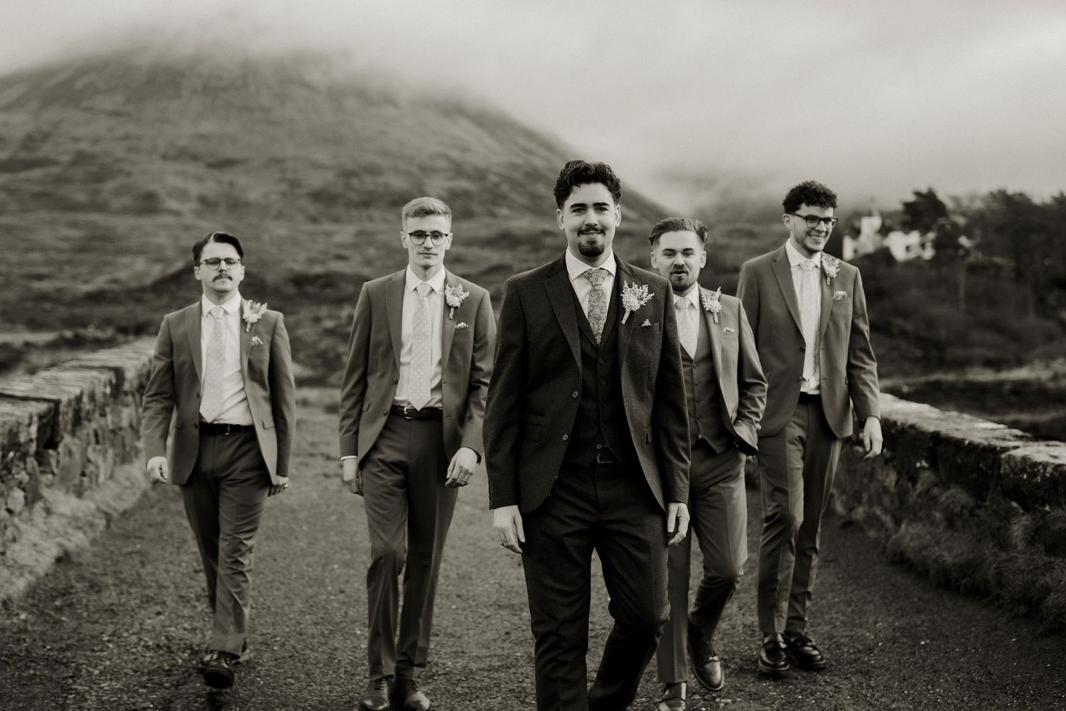 Black and white photo of groom leading groomsmen in suits walking on Scottish highland bridge.