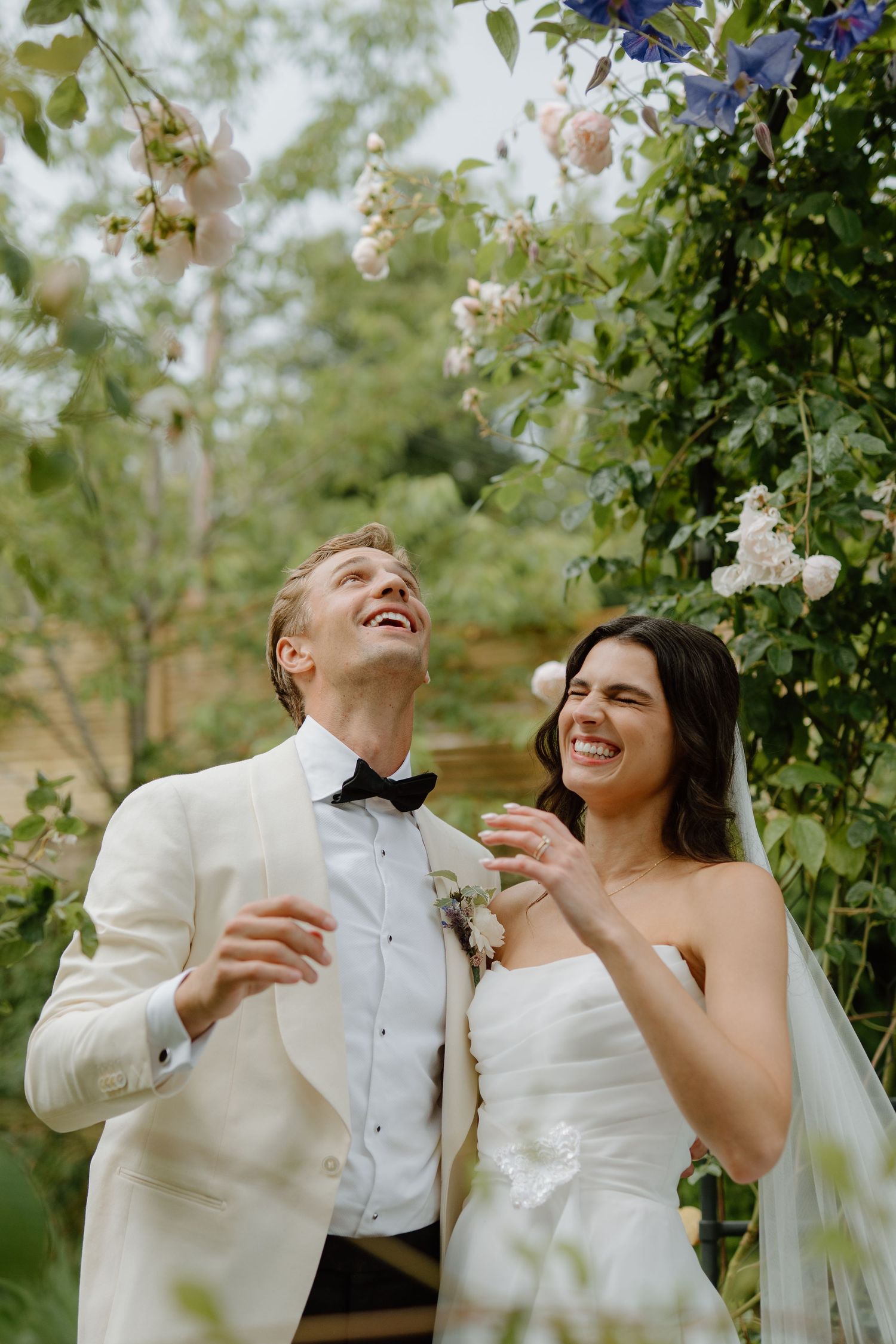 Laughing bride and groom stand beneath a flowering garden arch, both joyfully looking up at the blossoms.