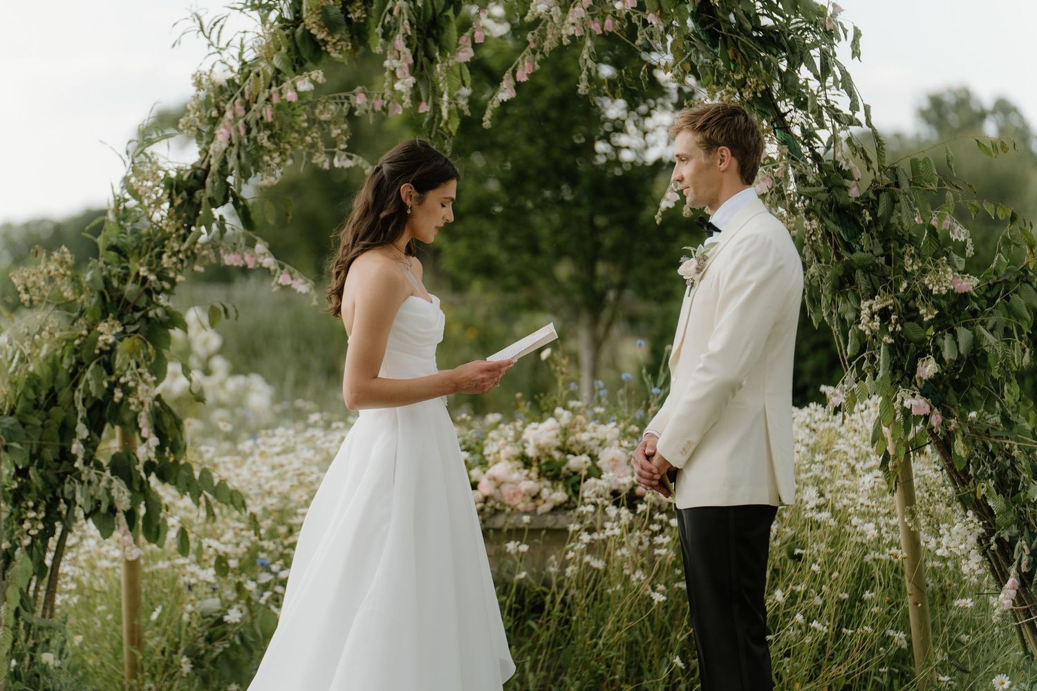 Bride and groom exchanging vows under lush greenery arch in outdoor wildflower garden ceremony.