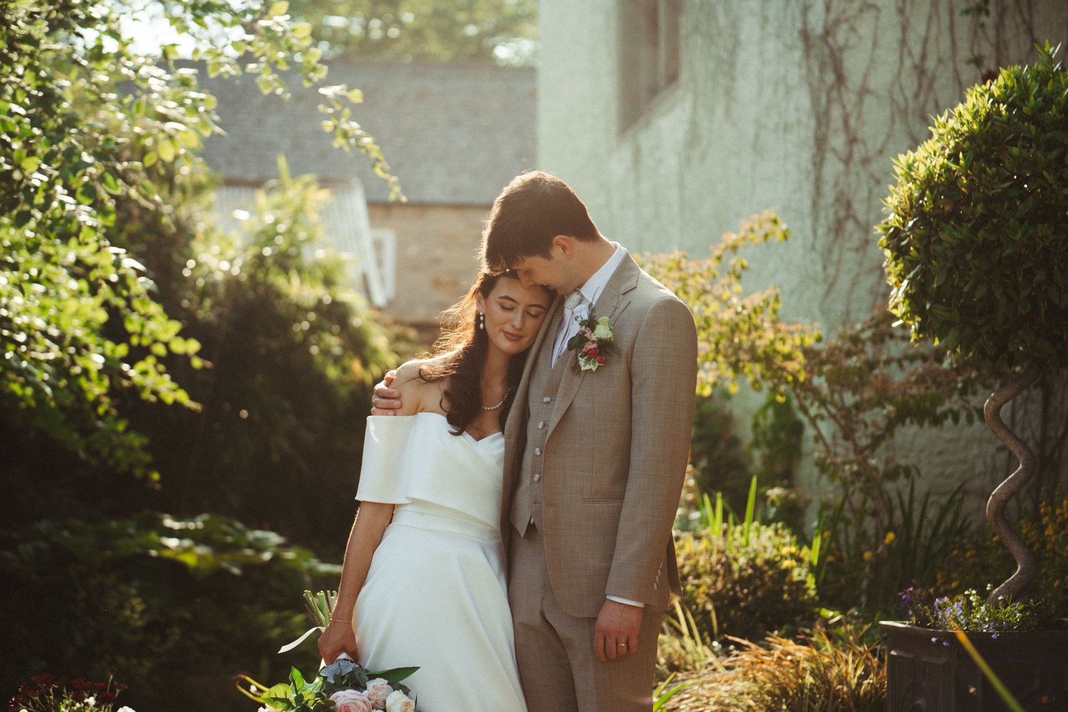 Bride and groom sharing tender embrace in sunlit garden, groom in beige suit with boutonniere.