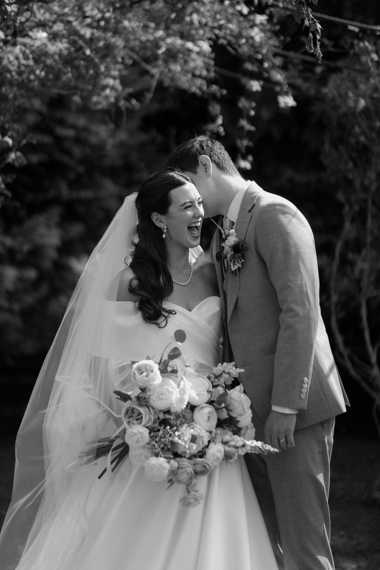Black and white photo of joyful bride and groom laughing together beneath flowering garden trees.