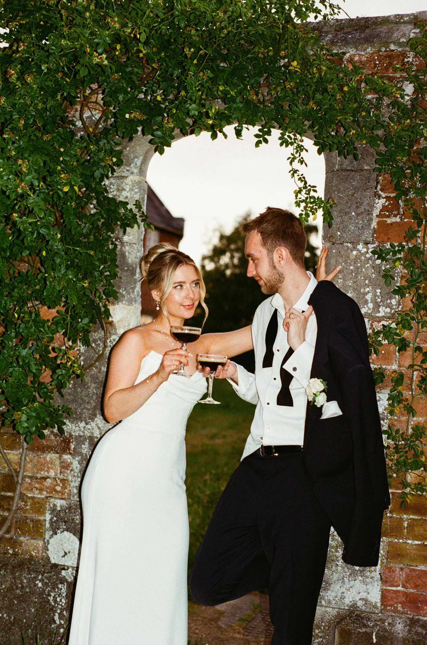Bride and groom in evening attire toast with red wine beneath ivy-covered stone archway at dusk.
