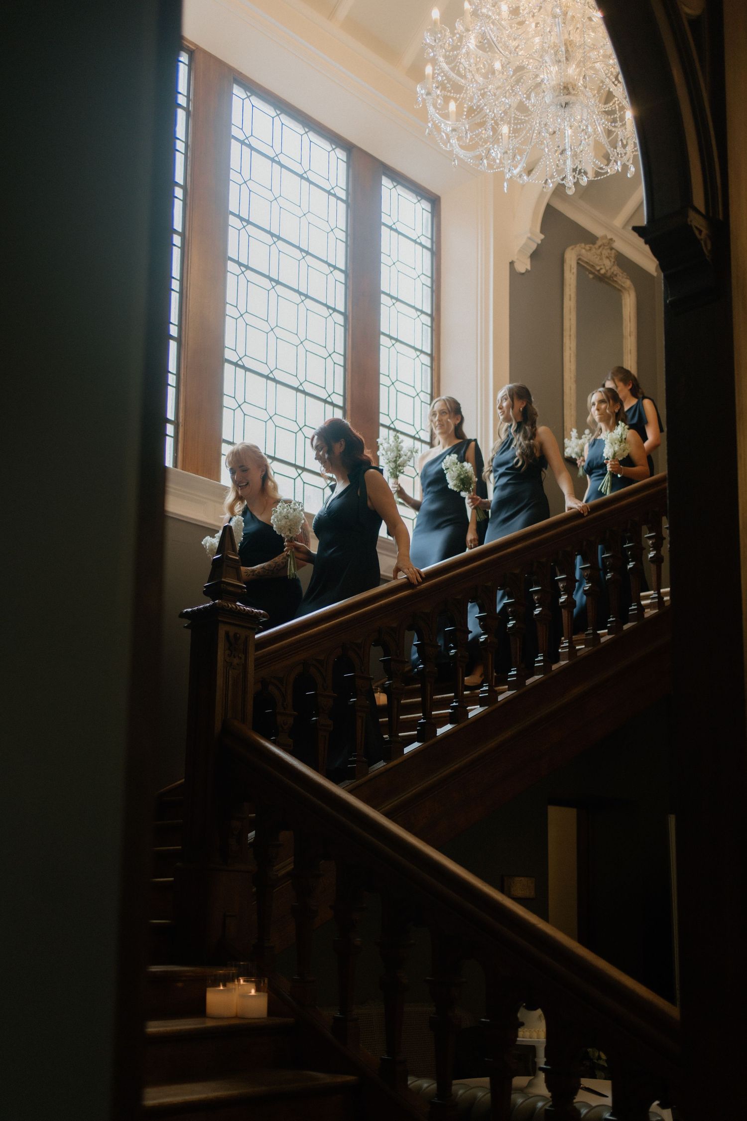 Bridesmaids in teal gowns descending a grand wooden staircase beneath a crystal chandelier at a wedding.