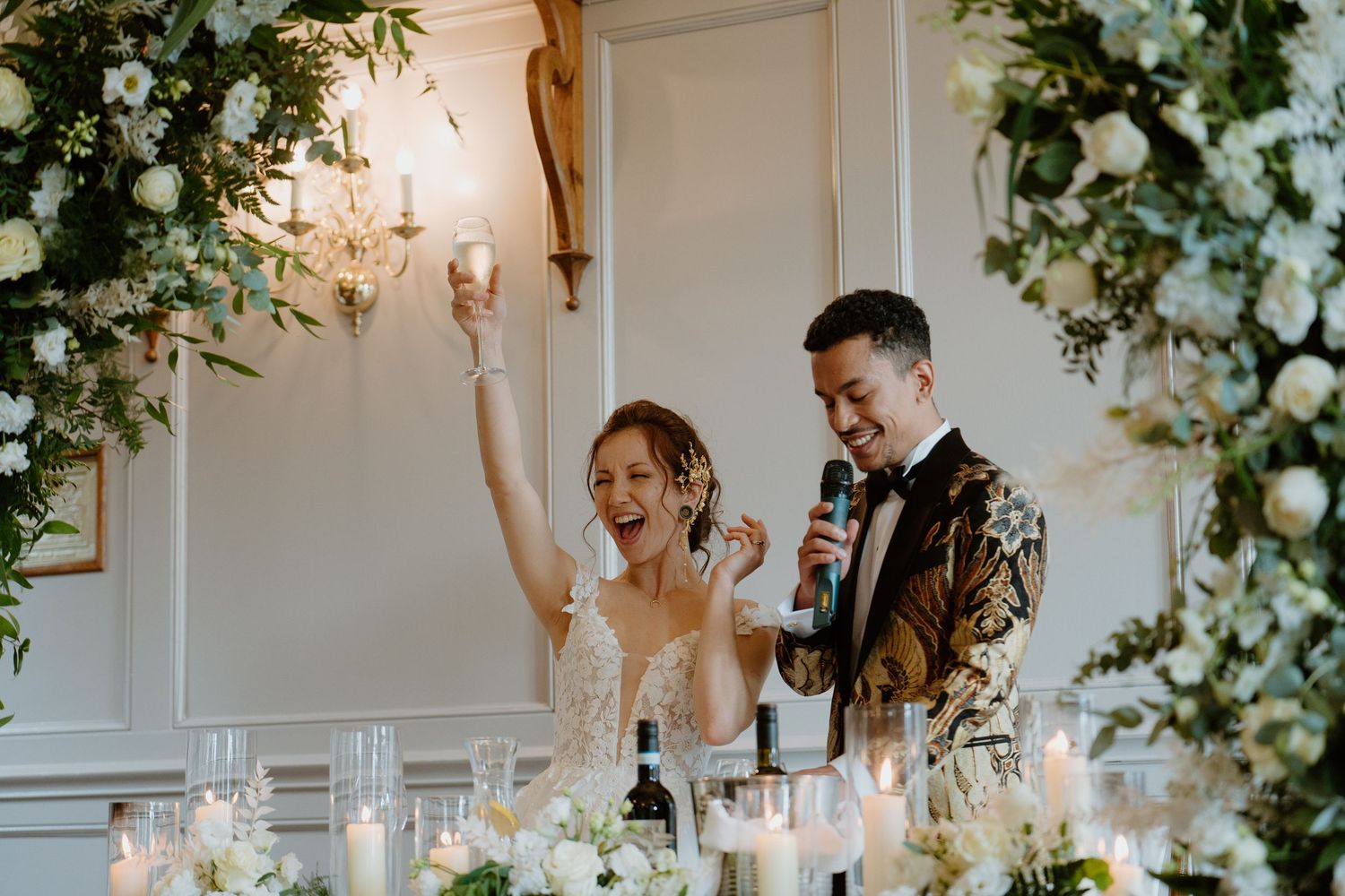 Bride and groom celebrating at wedding reception head table surrounded by white flowers and candles.