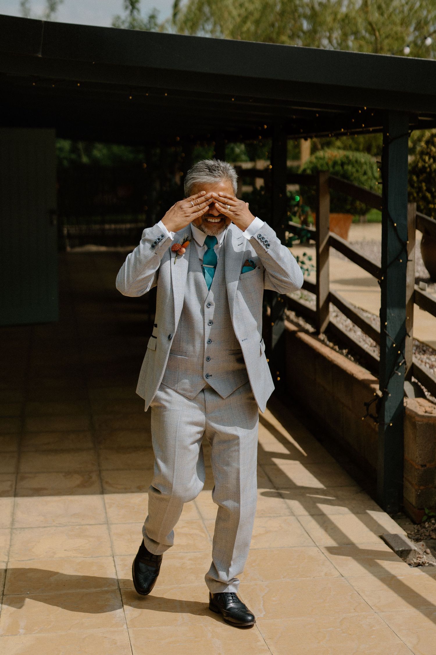 Groom in light grey three-piece suit covering his eyes during a first look wedding moment outdoors.