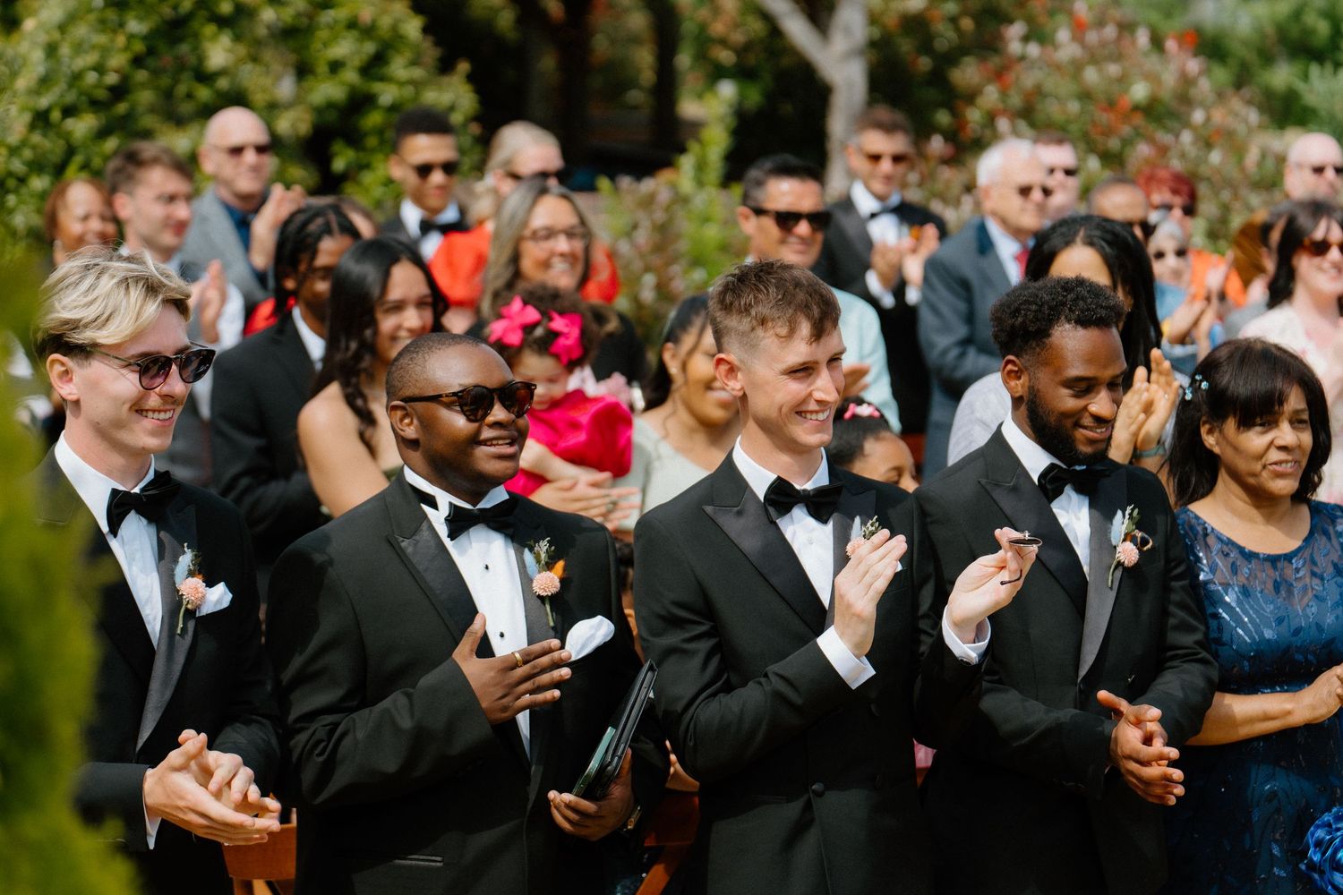 Groomsmen in black tuxedos clap and celebrate during outdoor wedding ceremony surrounded by joyful guests.