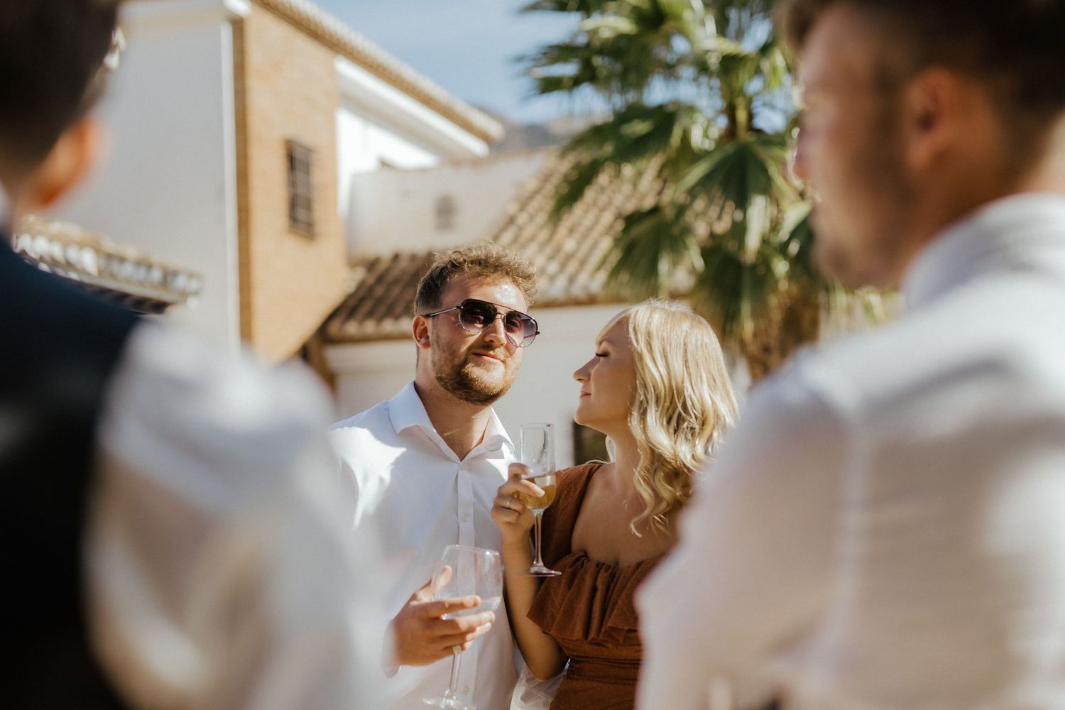 Wedding guests socializing outdoors with champagne in sunny Mediterranean setting.