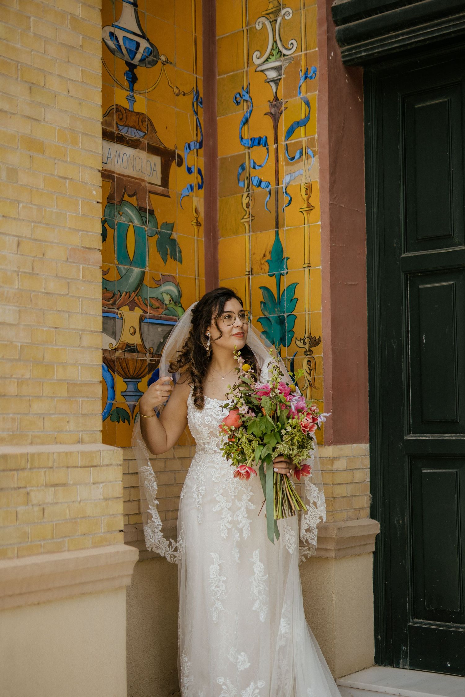 Bride in lace wedding gown holds colorful bouquet beside ornate tiled wall at venue.
