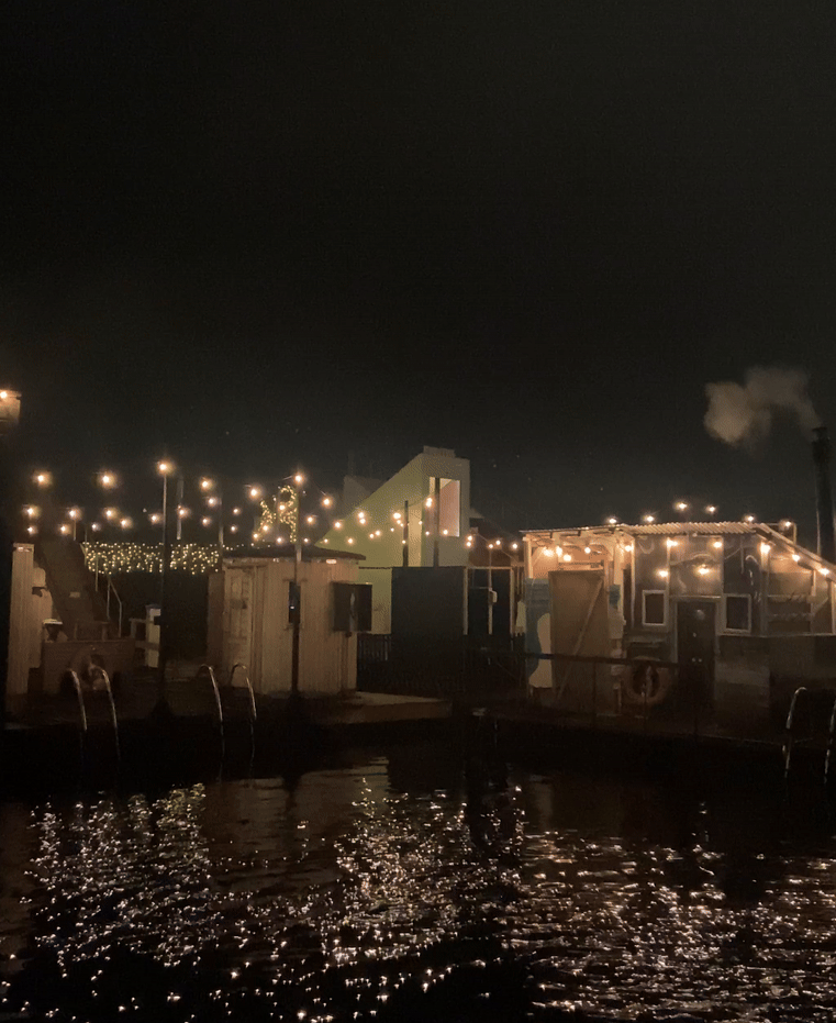 String lights illuminate a waterfront restaurant deck with reflections glimmering on the dark water at night.
