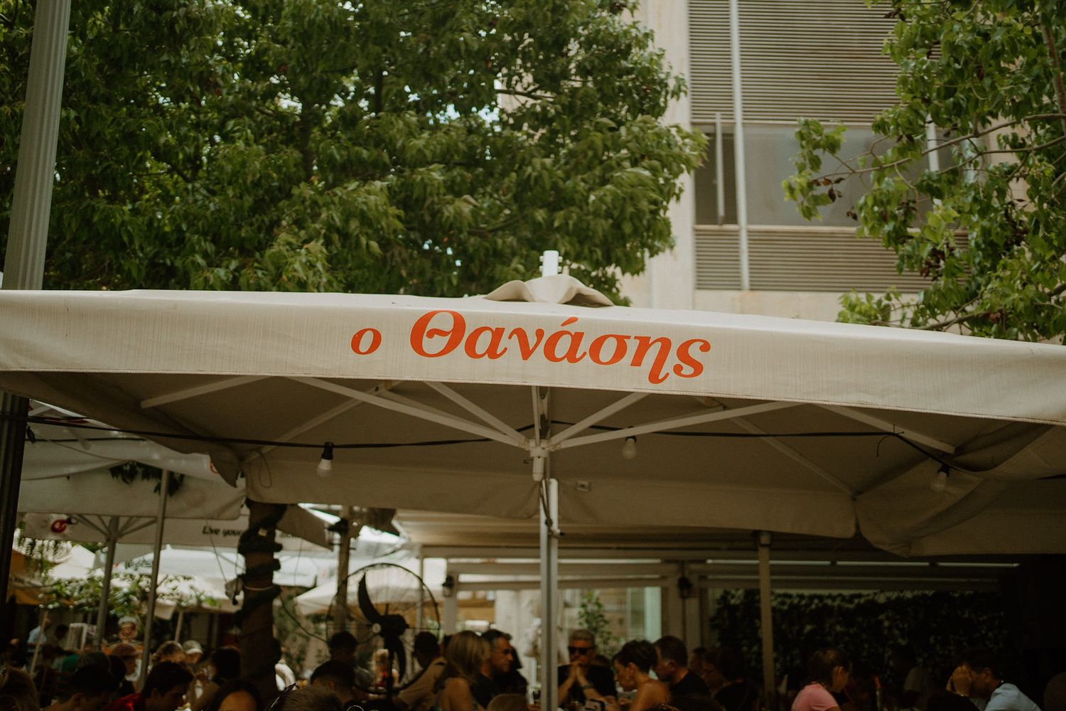 White restaurant umbrella with 'O Thanasis' text at an outdoor dining area surrounded by trees and patrons.