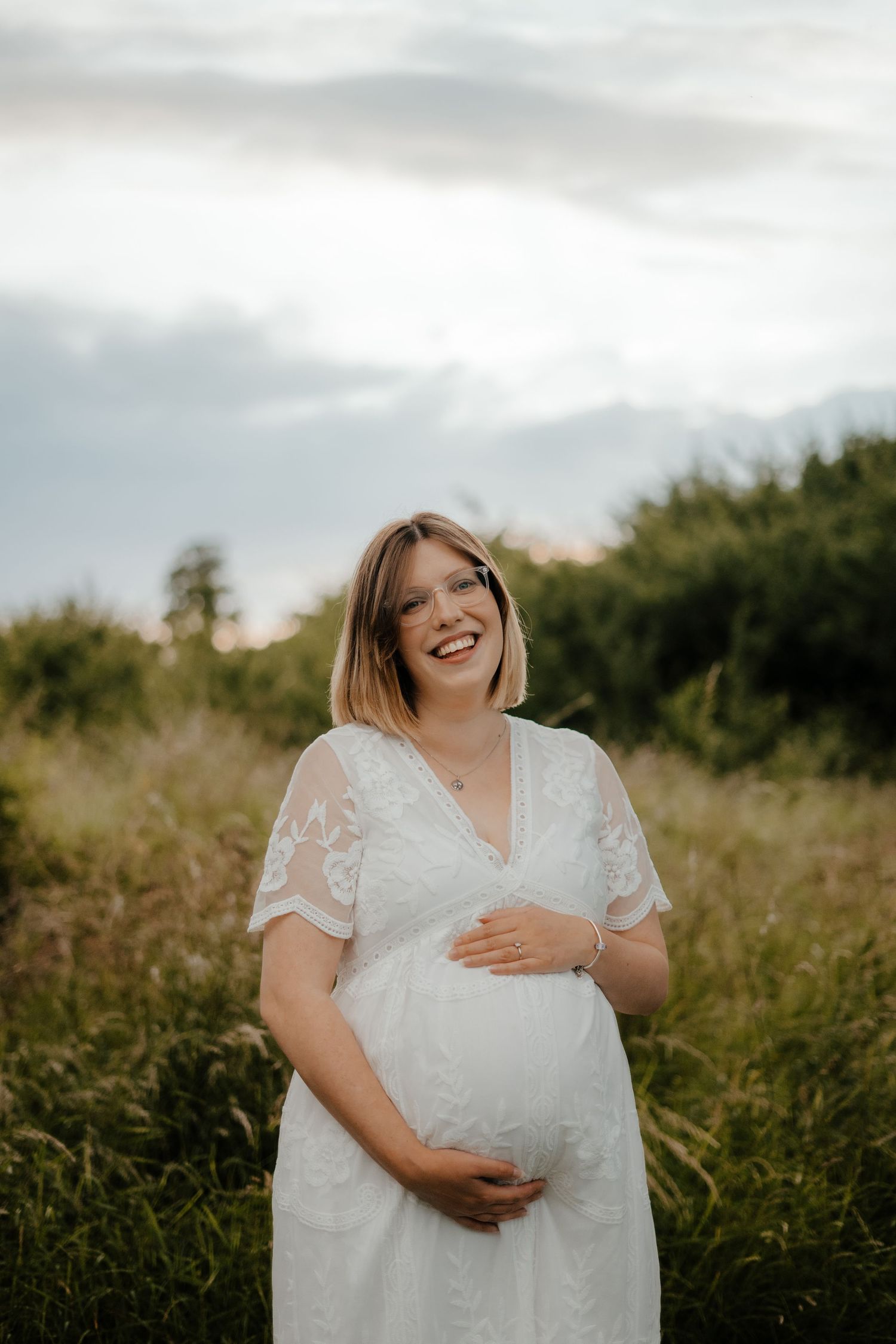 A happy pregnant woman in a white lace dress holds her baby bump in a meadow.