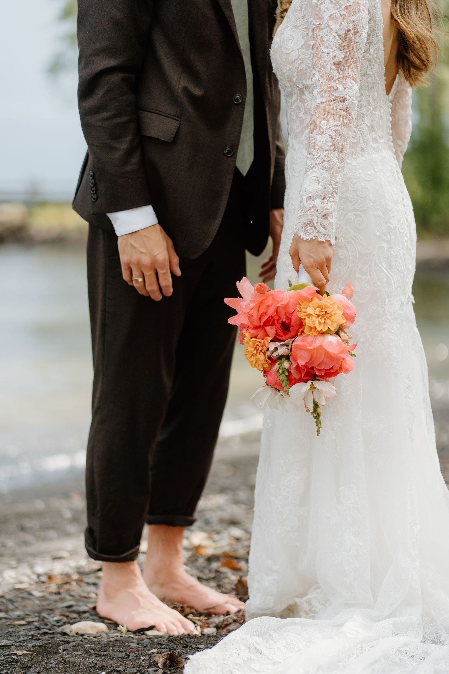 Close-up of bride holding vibrant coral and orange bouquet beside barefoot groom on rocky shore.
