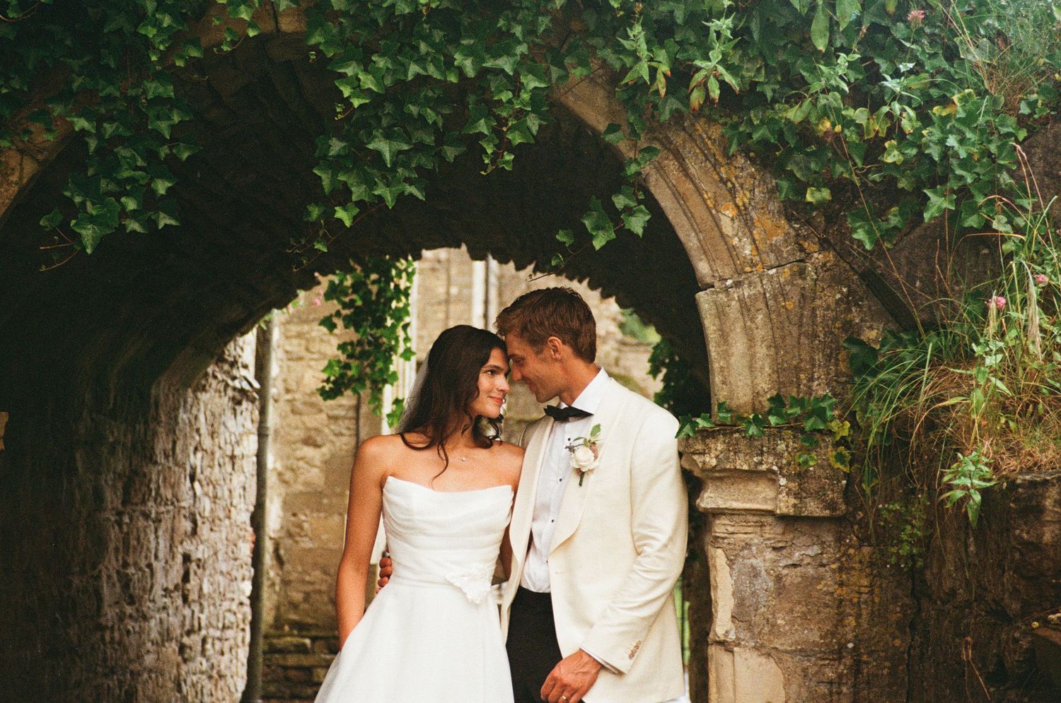 Bride and groom sharing intimate moment under ivy-covered stone archway.