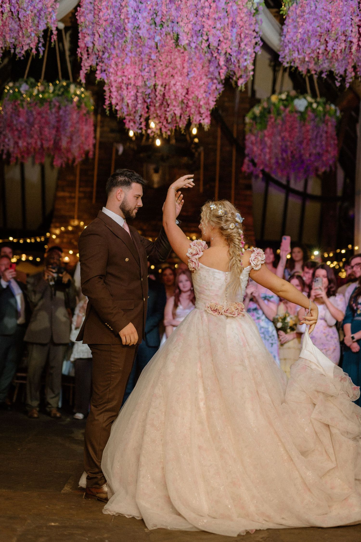 Bride and groom share first dance beneath stunning hanging purple wisteria in a warmly lit barn venue.