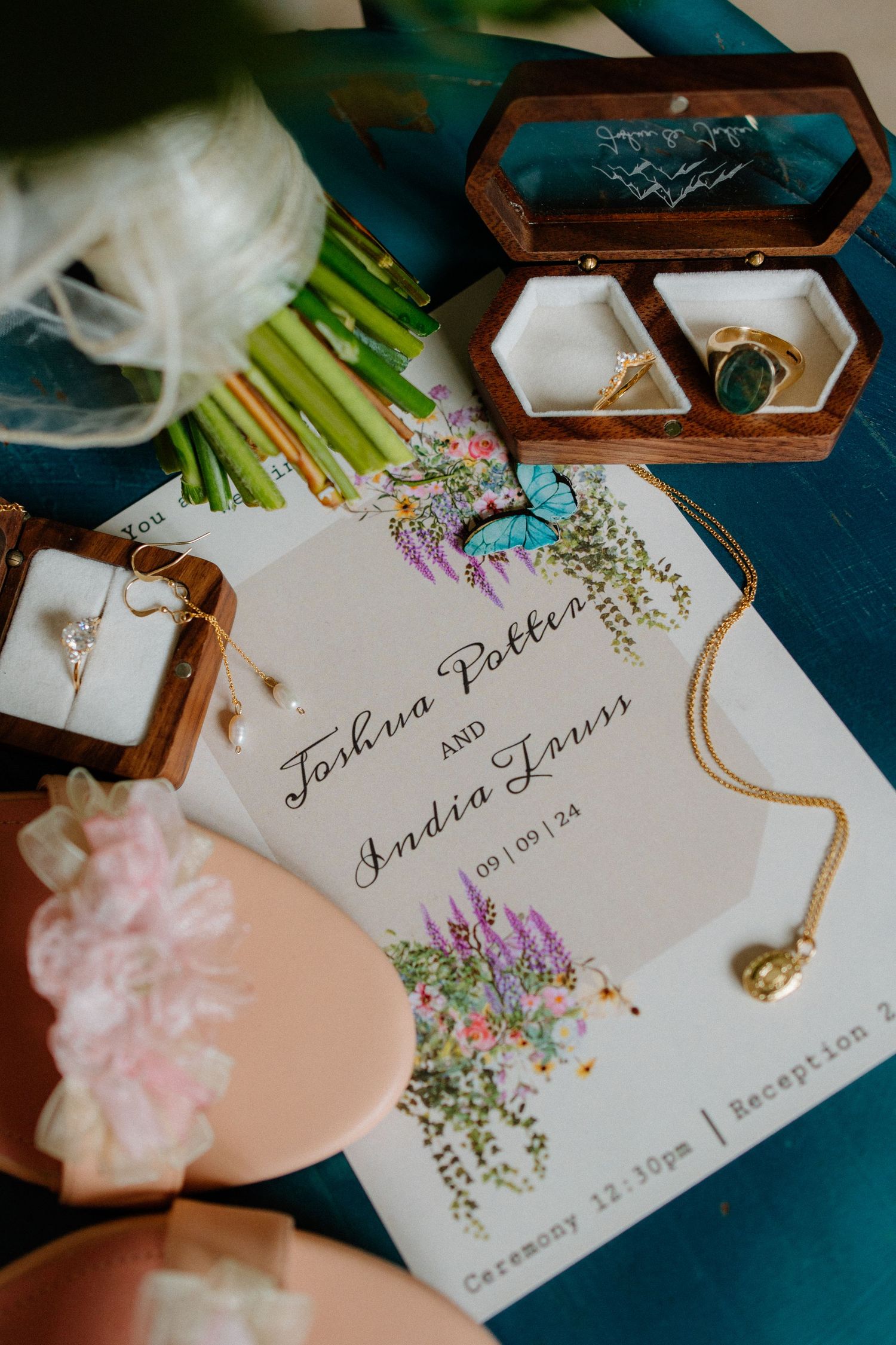 Wedding flat lay featuring floral invitation for Joshua Potter and Andia Louis with gold jewelry and pink shoes.